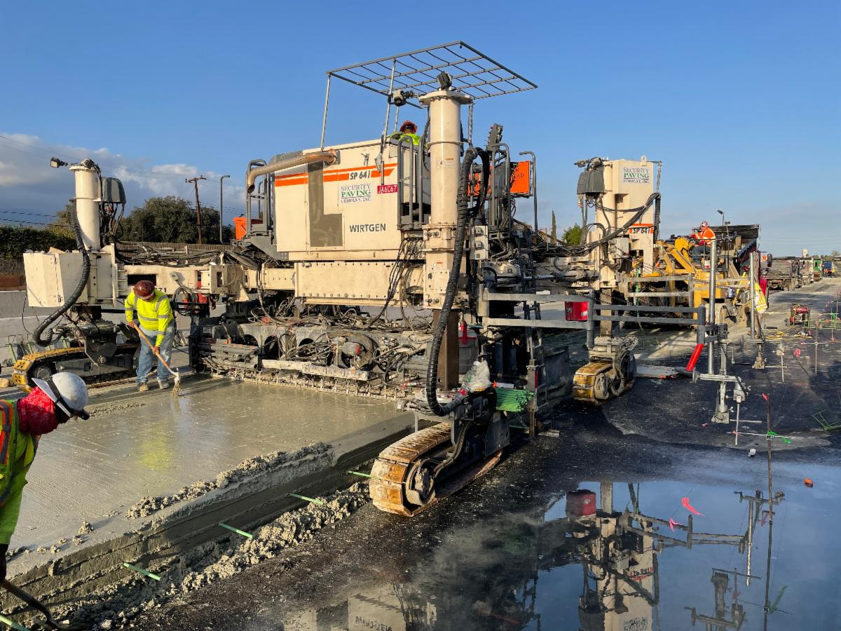 Construction workers operate heavy machinery to lay and smooth wet concrete on a road under clear skies.