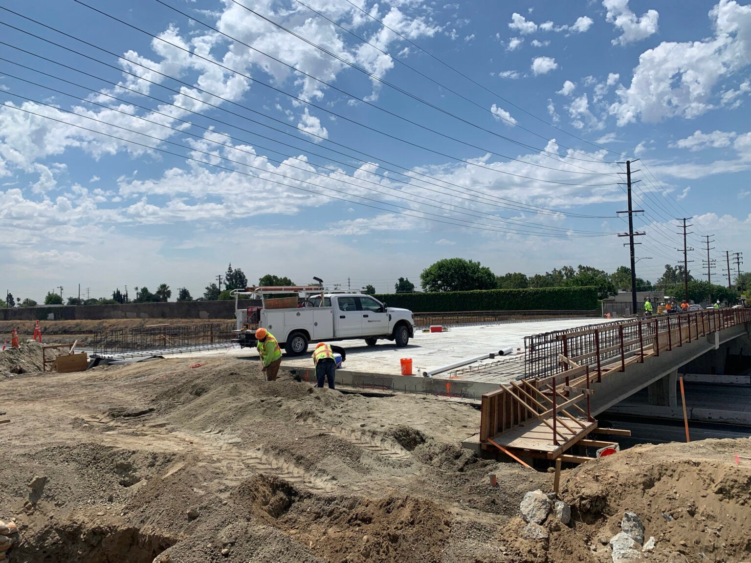 Construction workers and a white utility truck at a bridge construction site under a partly cloudy sky, with power lines visible overhead.