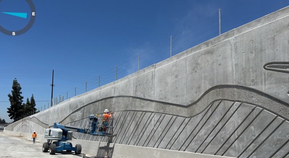 Two construction workers on a lift work on a large, textured concrete retaining wall under a clear blue sky. A compass is visible in the top left corner.