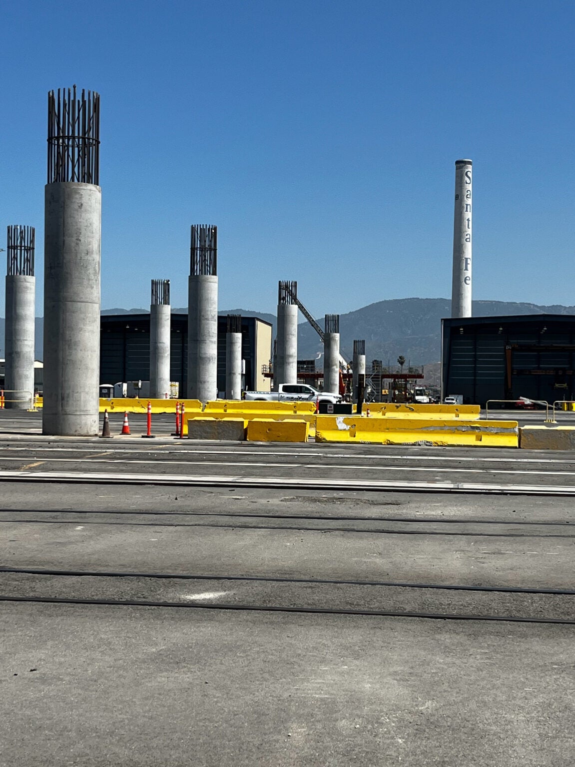 Rows of unfinished concrete columns stand on a construction site with yellow barriers and traffic cones under a clear blue sky; "Santa Fe" is visible on a tall white structure in the background.