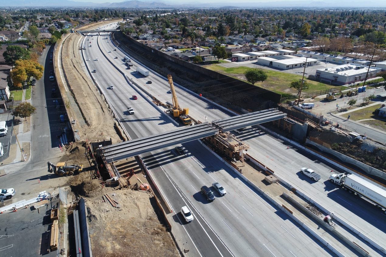 Aerial view of highway construction with cranes installing beams for a new overpass; vehicles travel on both sides of the work zone.