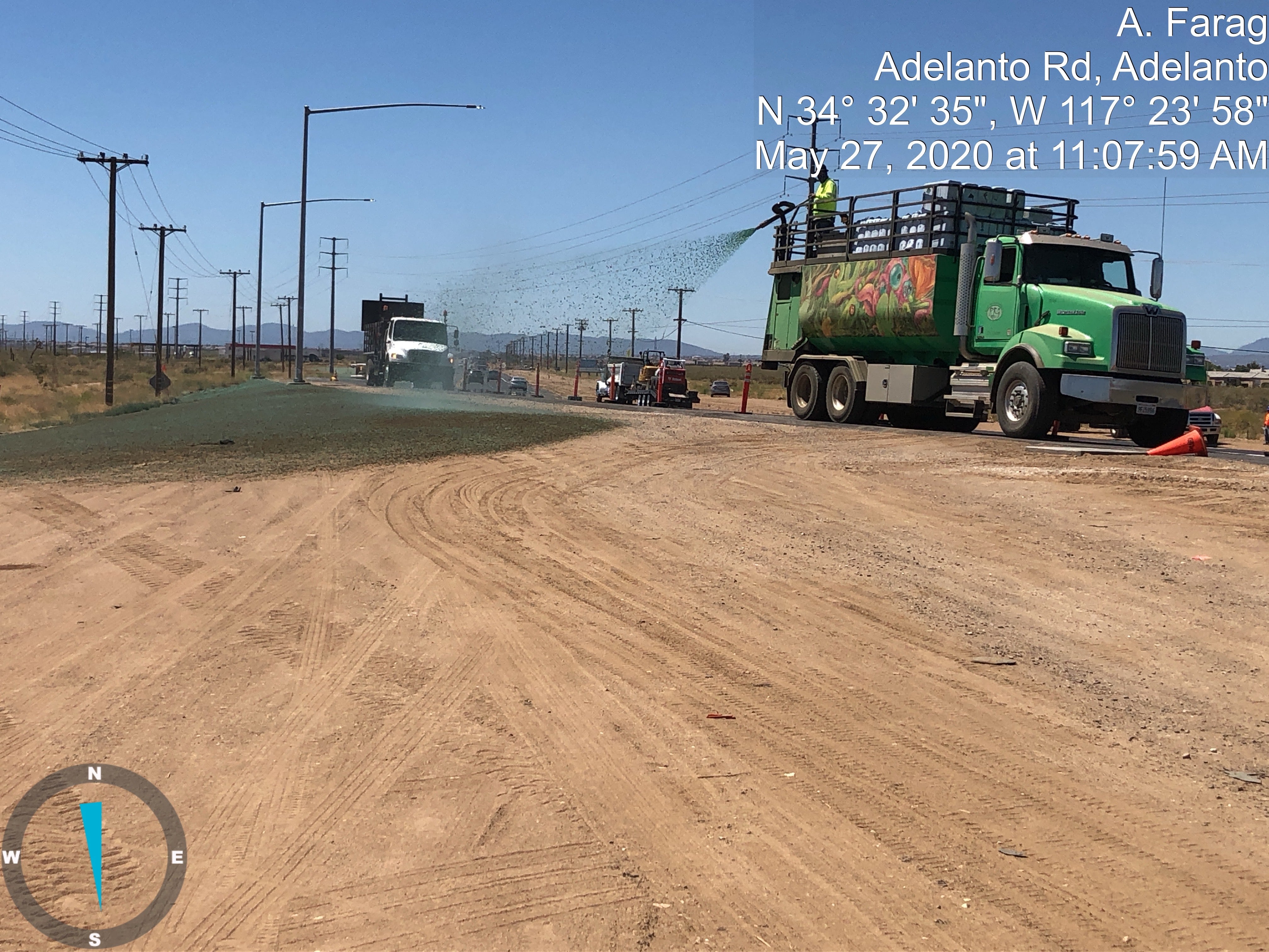 A green truck sprays material onto a dirt road under clear skies while workers stand nearby; timestamp and location details are overlaid on the image.