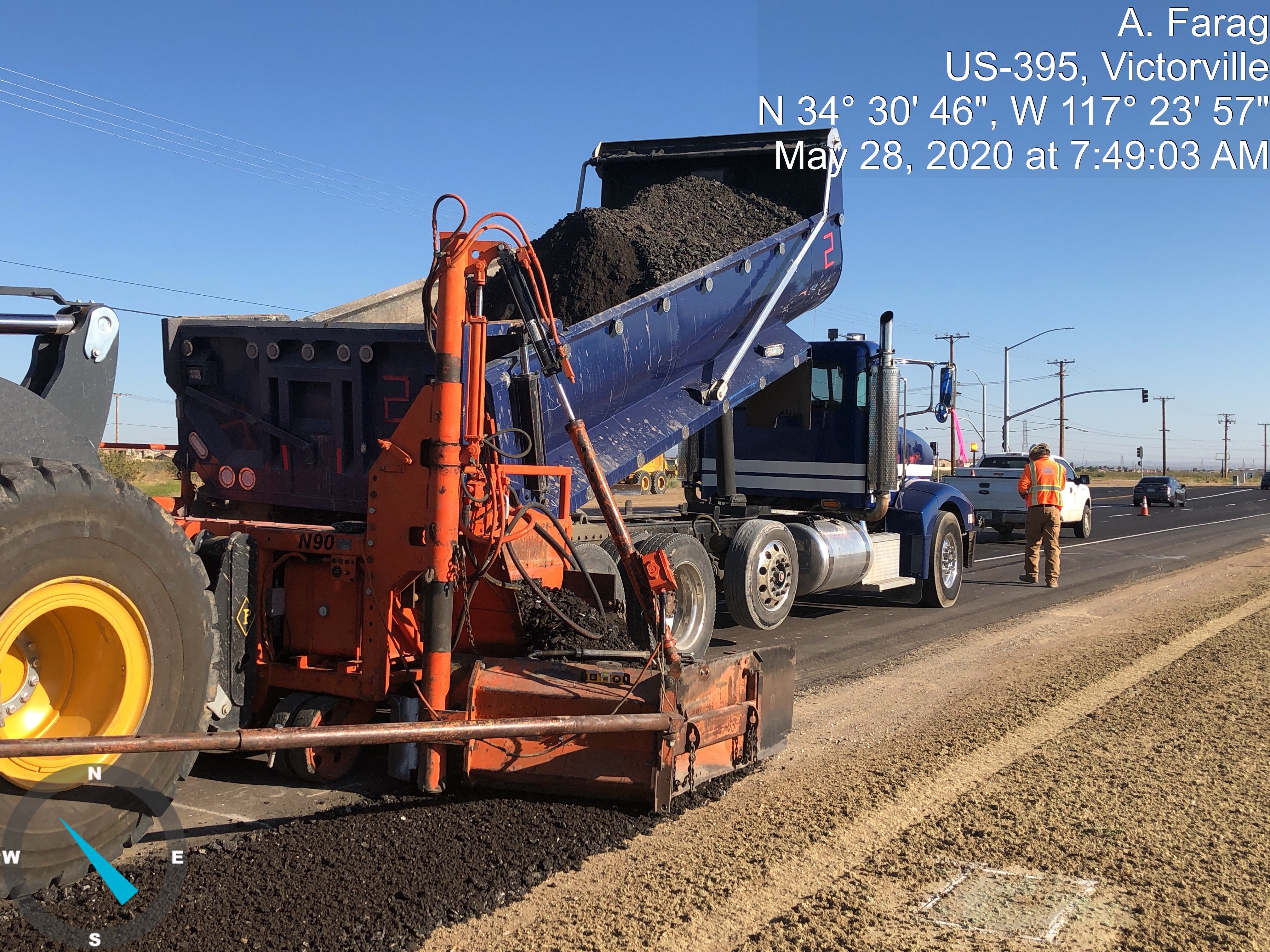 A dump truck unloads asphalt into a paving machine as workers resurface a highway under clear skies in Victorville, California.