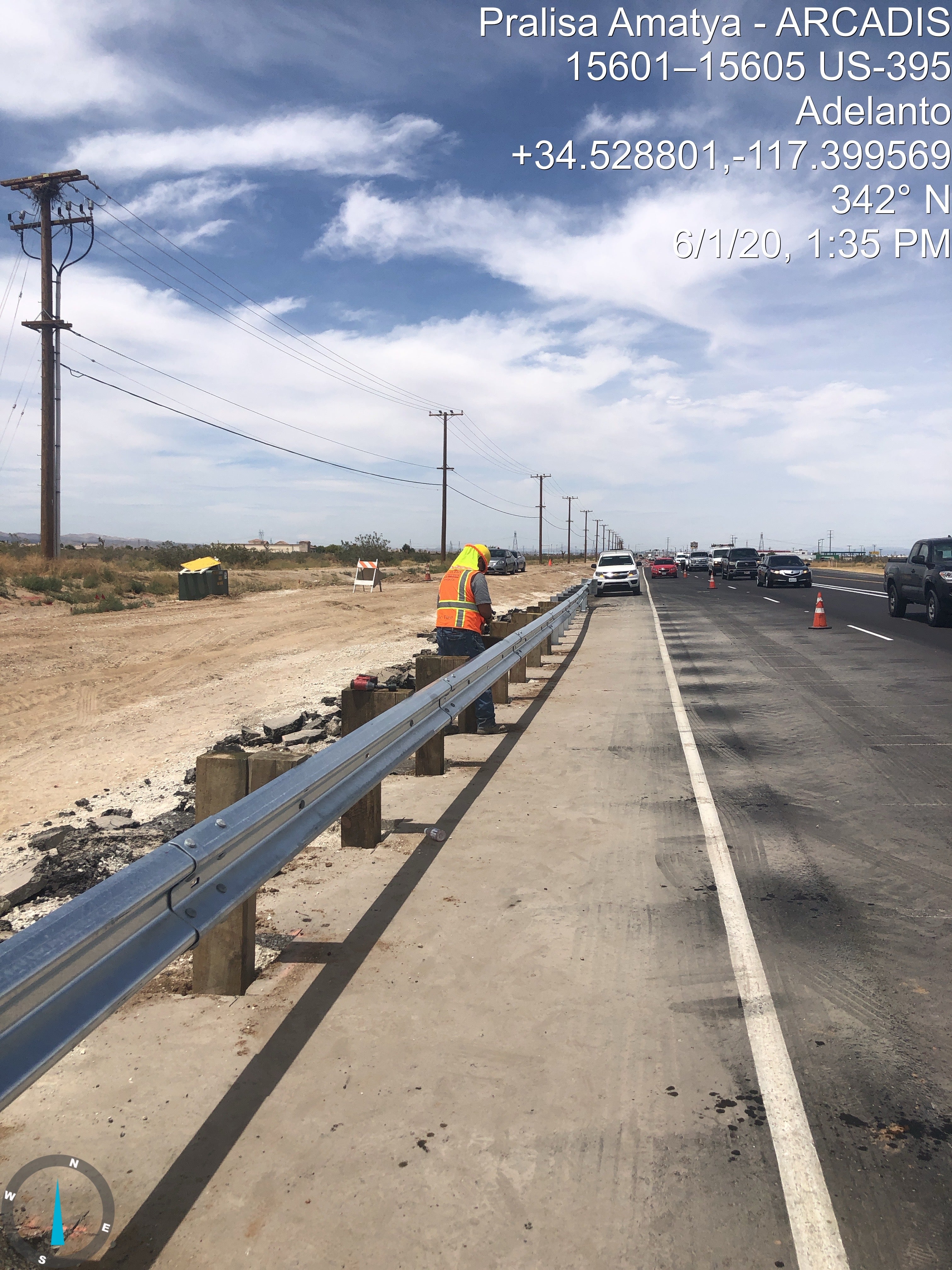 A road worker in a safety vest repairs a guardrail on the shoulder of a highway with traffic cones and vehicles nearby under a partly cloudy sky.