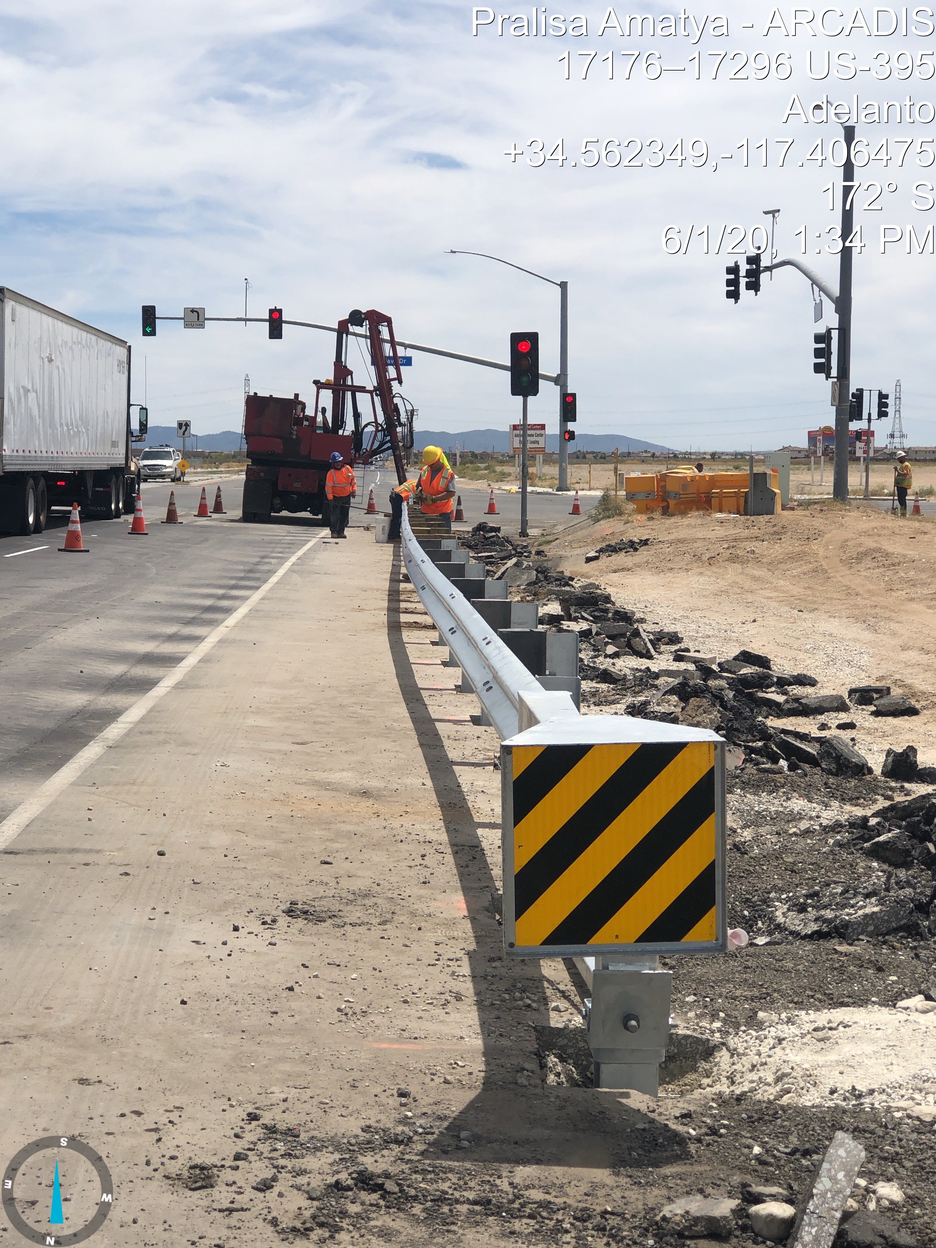 Road construction workers install a new guardrail along a partially closed lane; traffic cones and heavy machinery are present at the intersection.