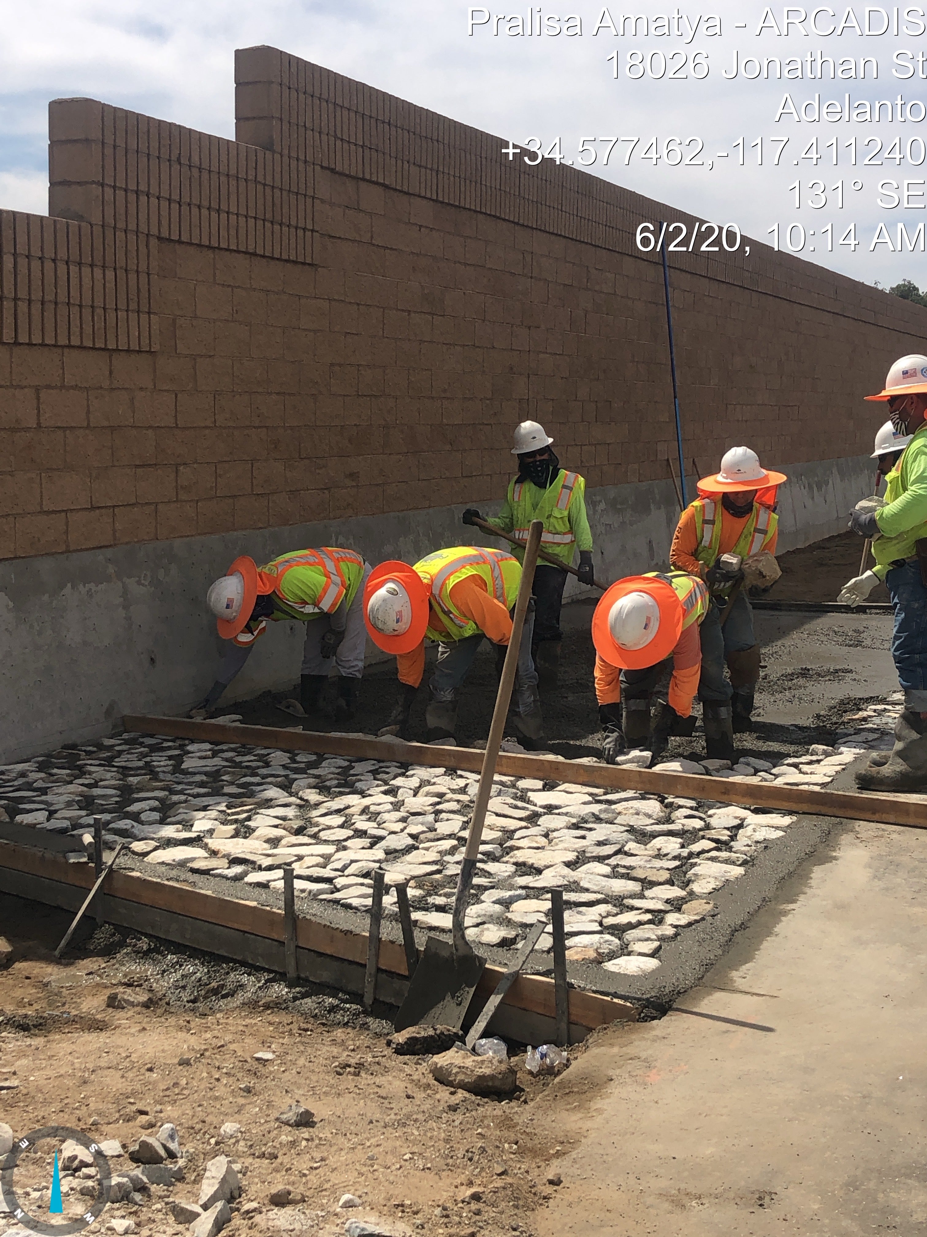 Six construction workers in safety gear lay and align stones on a concrete foundation beside a tall brick wall at a construction site.
