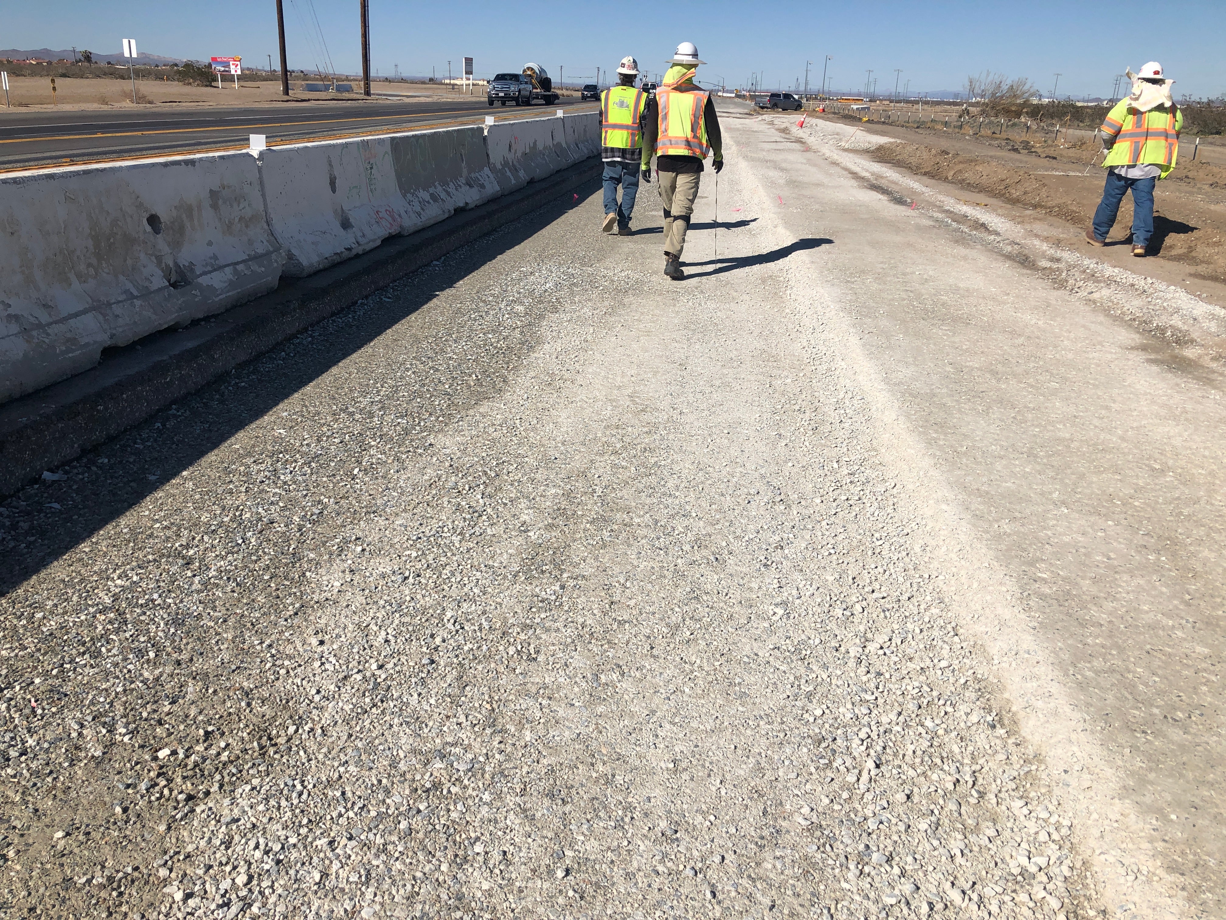 Three construction workers wearing safety vests and helmets walk on a gravel road next to concrete barriers at a road construction site.