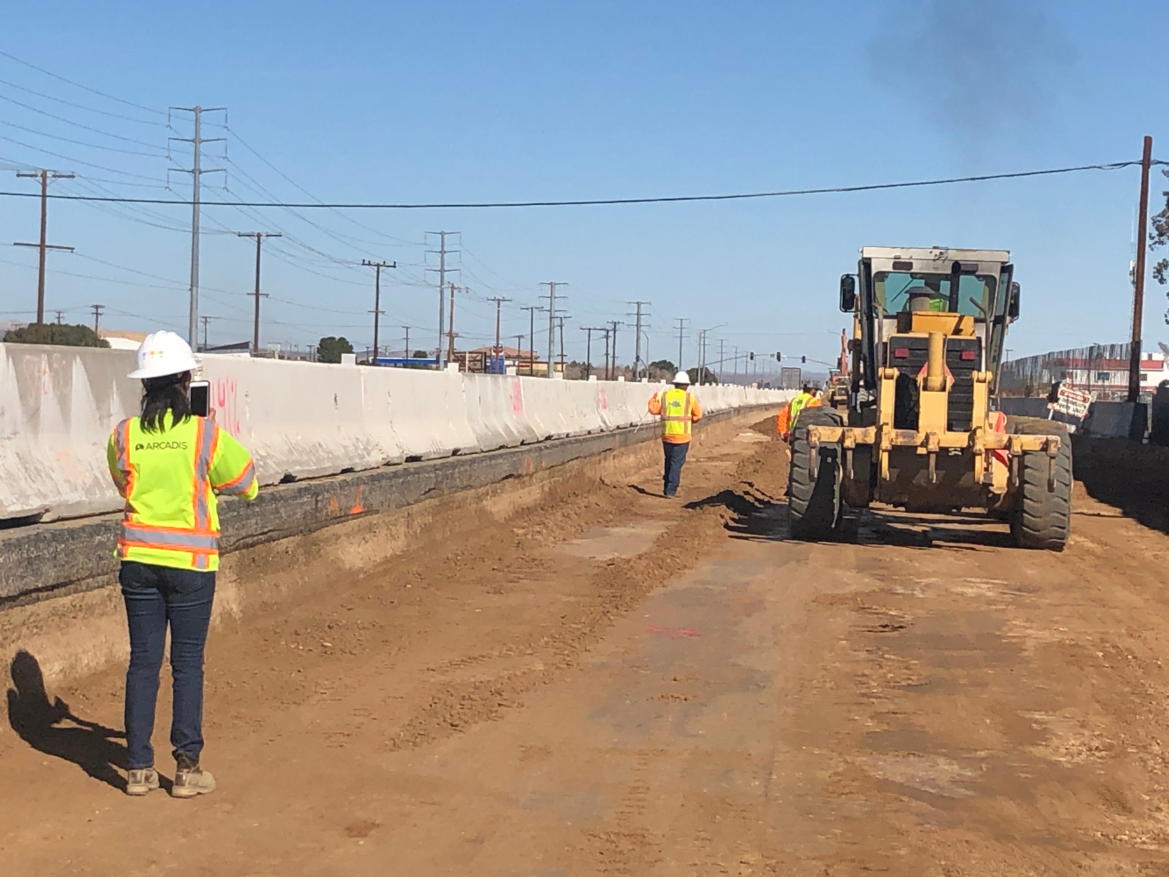 Construction workers in safety vests and helmets work on a dirt road next to concrete barriers; a large grader vehicle levels the ground.