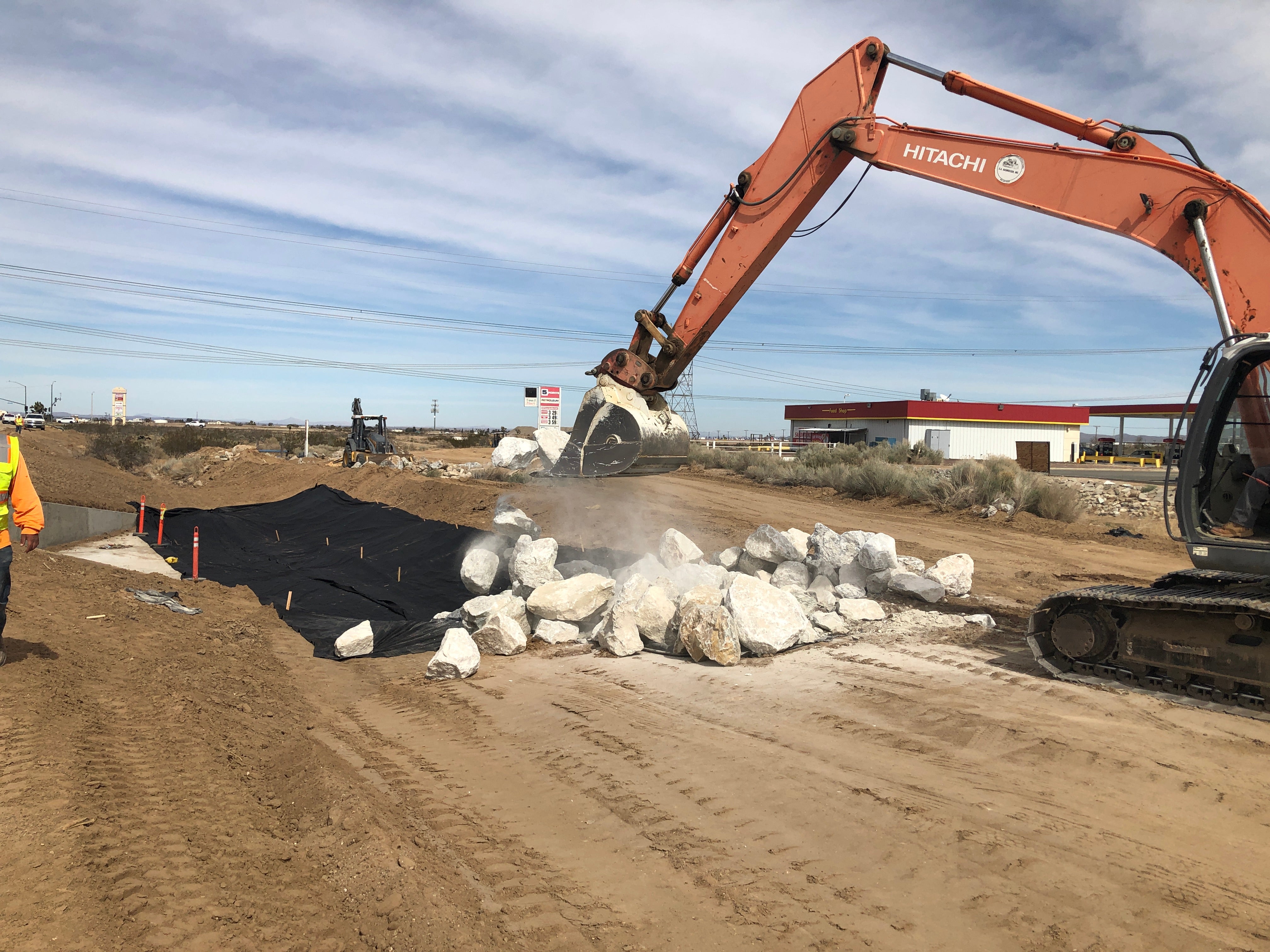 An excavator places large rocks onto black geotextile fabric on a dirt road construction site, with workers and industrial buildings visible in the background.