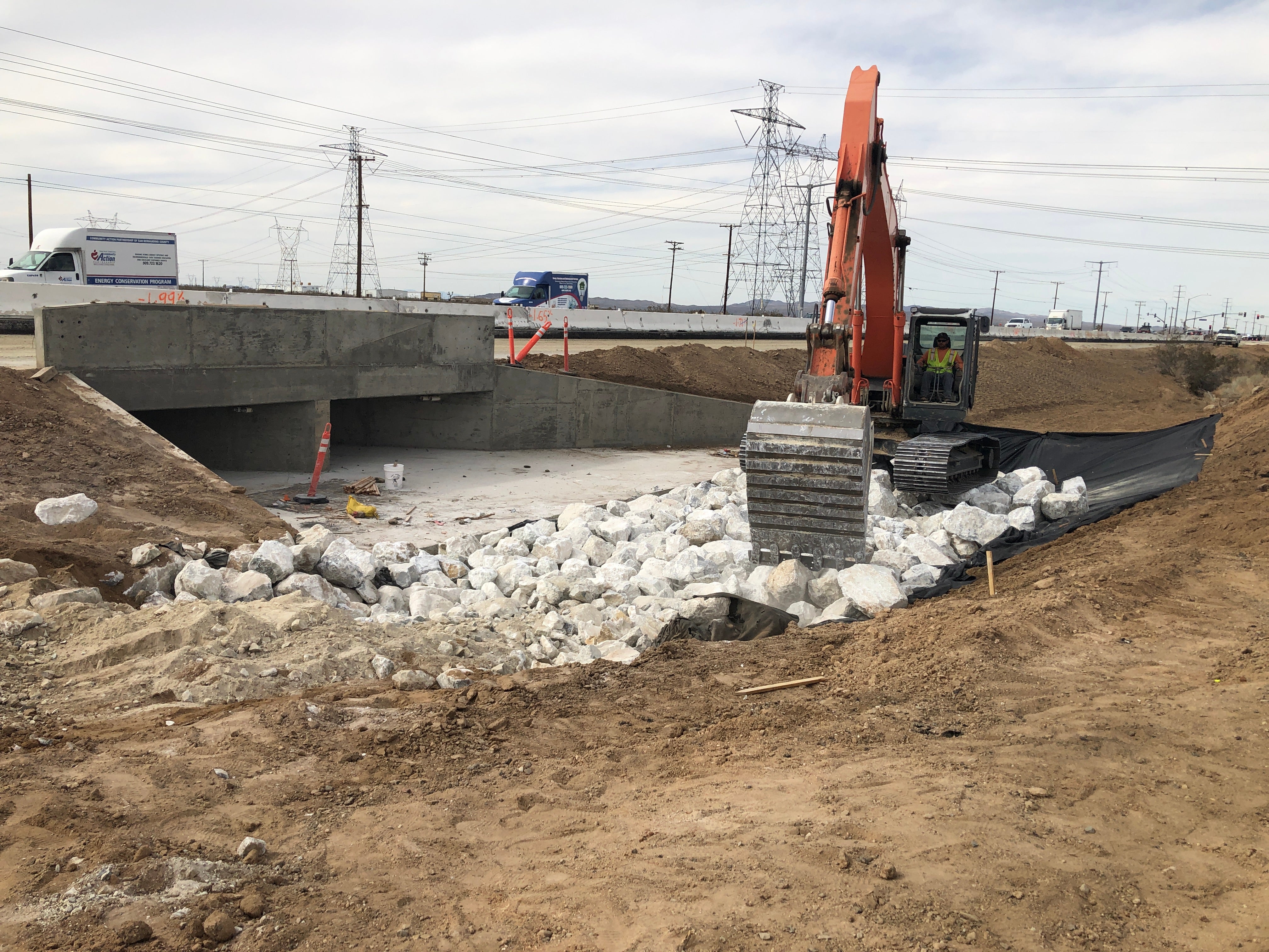 An excavator places rocks near a concrete culvert at a construction site by a highway, with vehicles and power lines in the background.