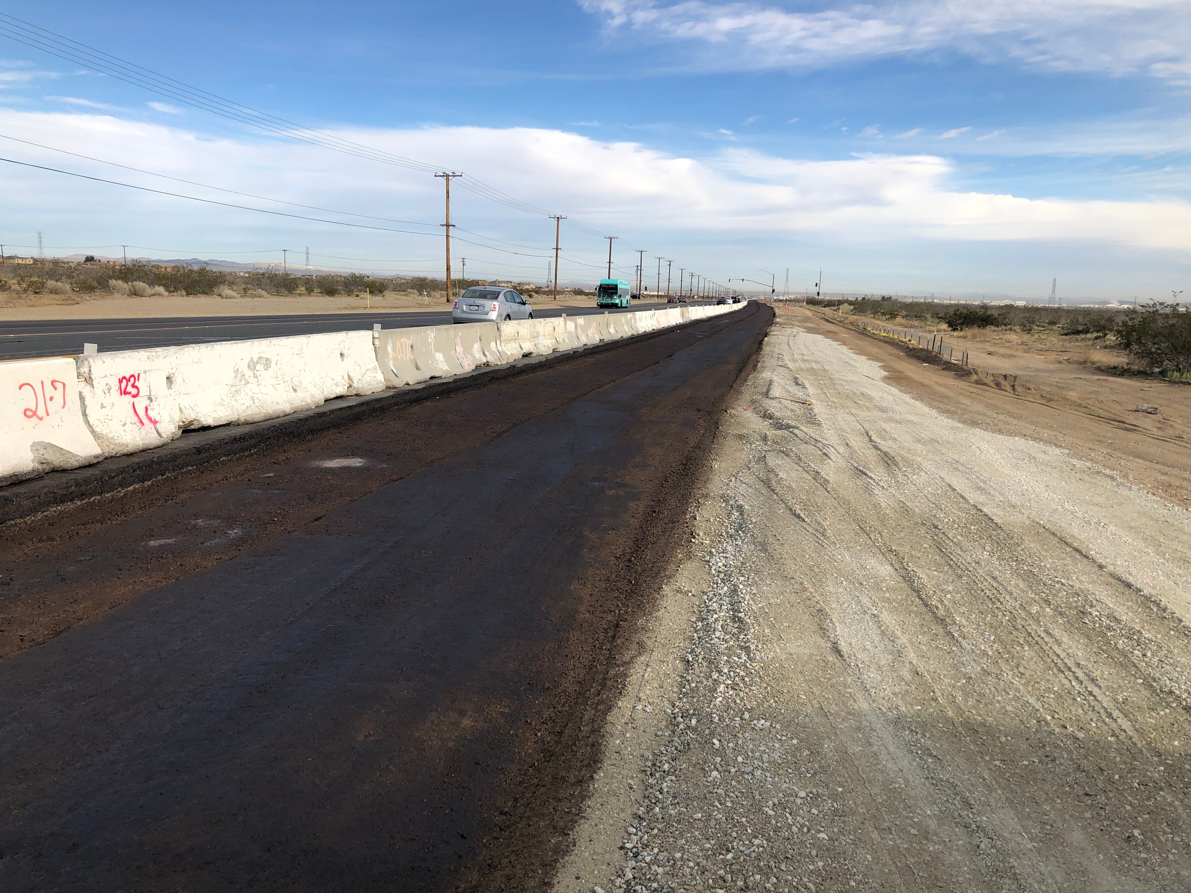 A newly paved road runs parallel to a concrete barrier and a gravel-covered area, with cars driving on the adjacent road in a desert landscape.