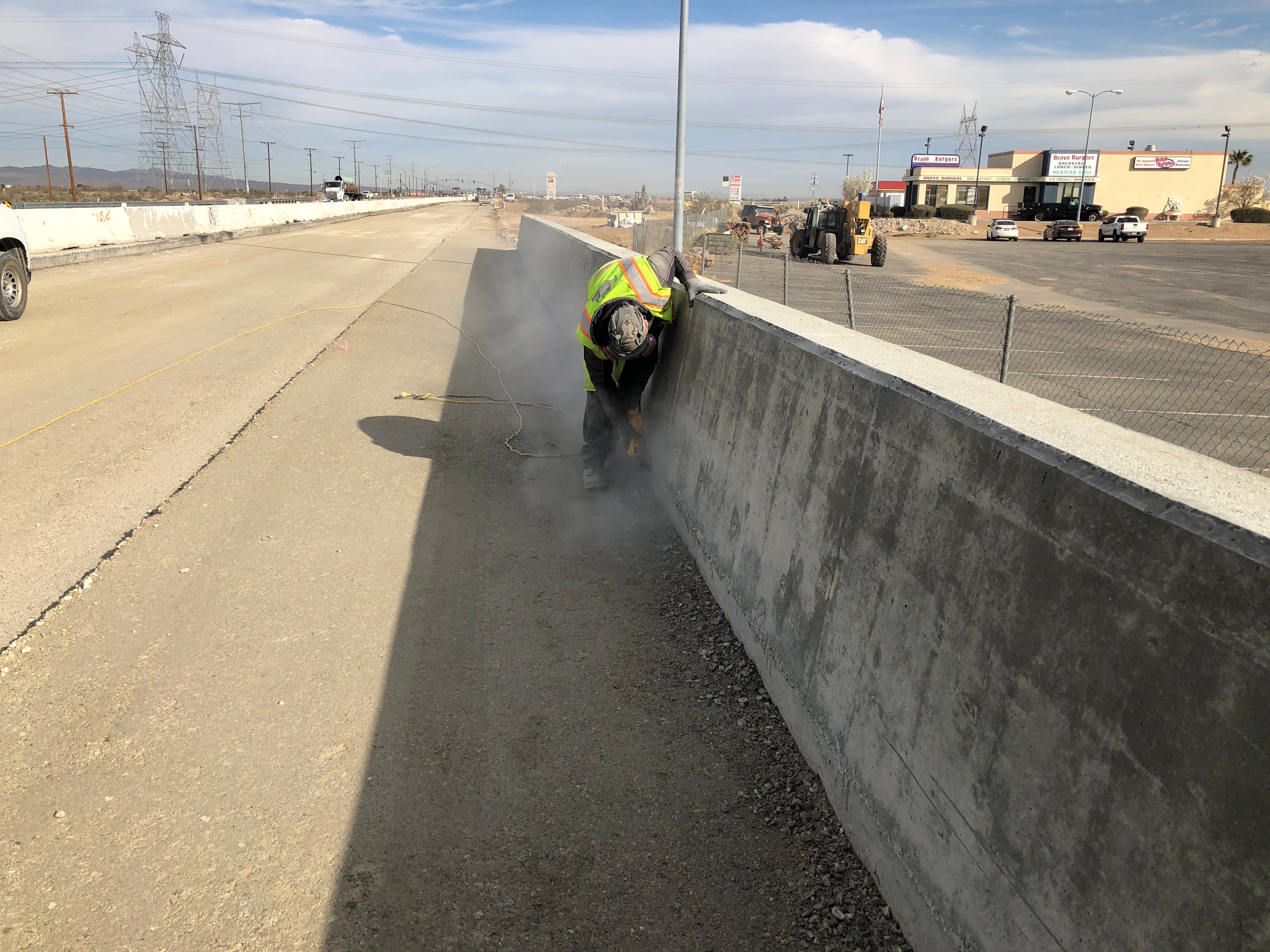 A construction worker in a safety vest uses power tools to work on a concrete barrier along a dusty highway under clear skies.