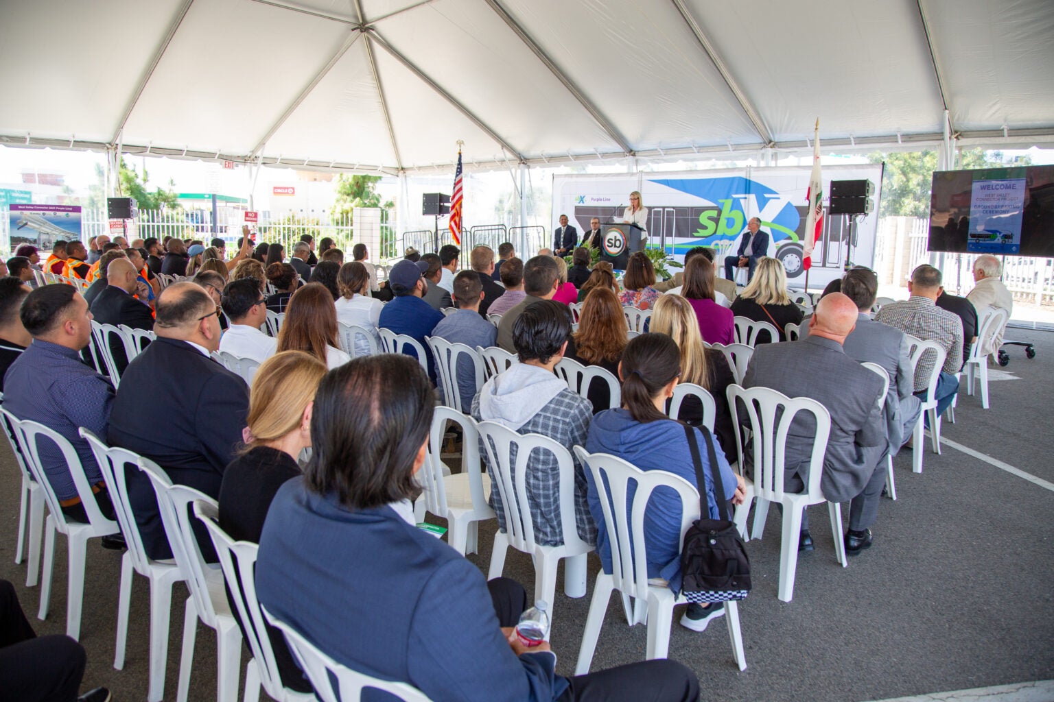 A large group of people seated under a white tent listens to a speaker at a podium during an outdoor event; flags and a transit bus are visible in the background.