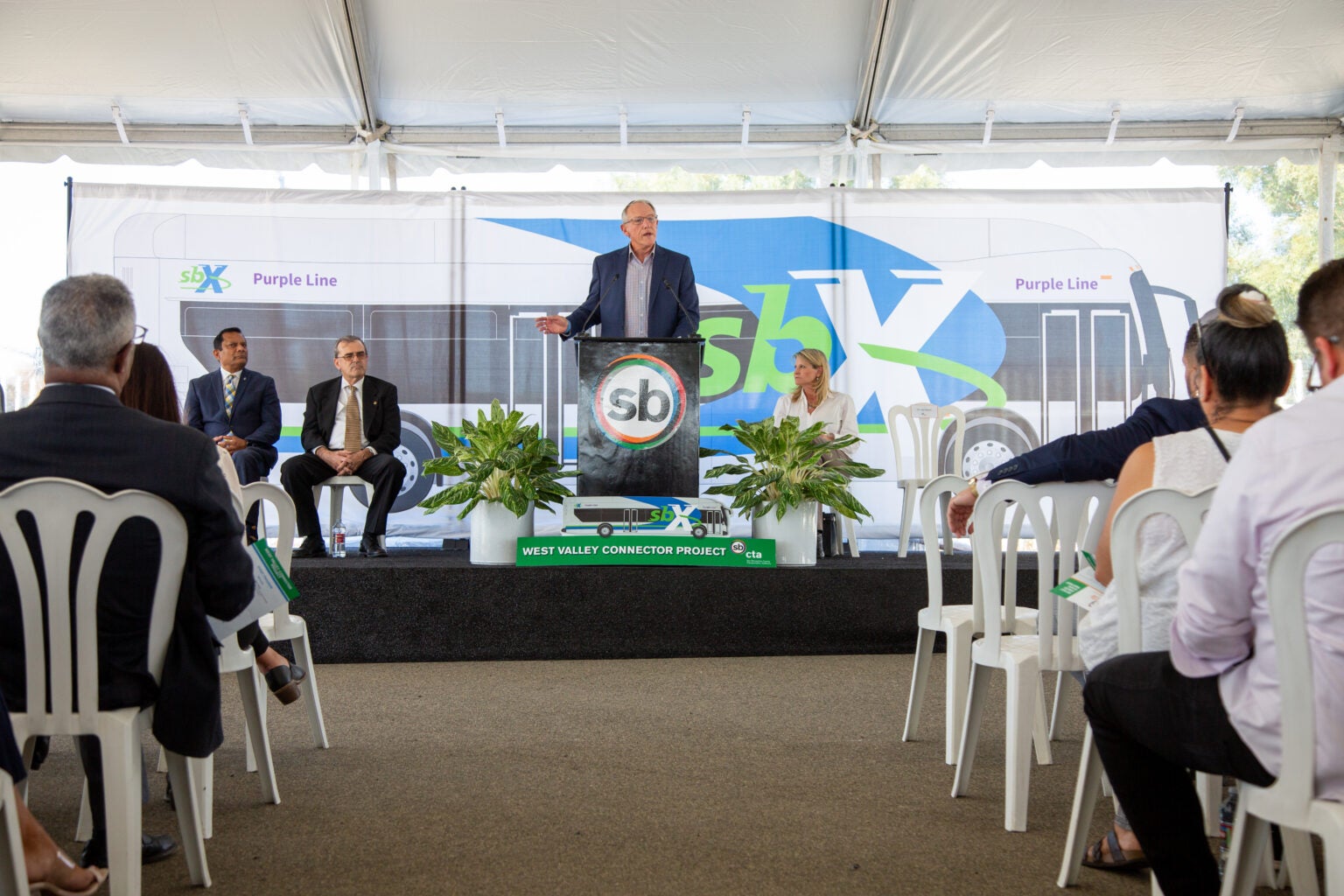 A man stands at a podium speaking to an audience at an event, with four people seated on stage and a bus graphic backdrop behind them.