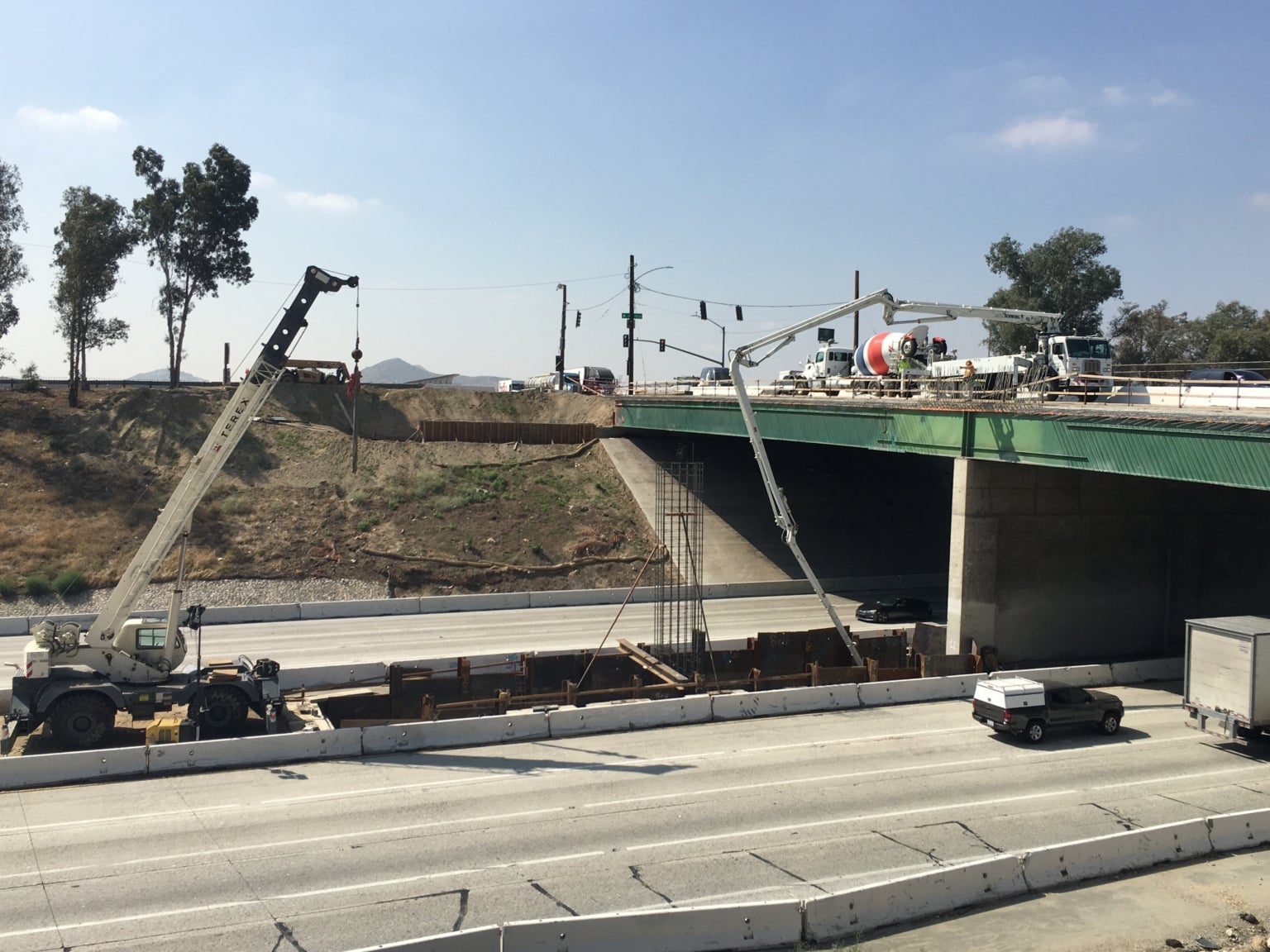 Construction cranes and concrete mixer working on a bridge above a multi-lane highway, with vehicles passing underneath and barriers separating traffic.