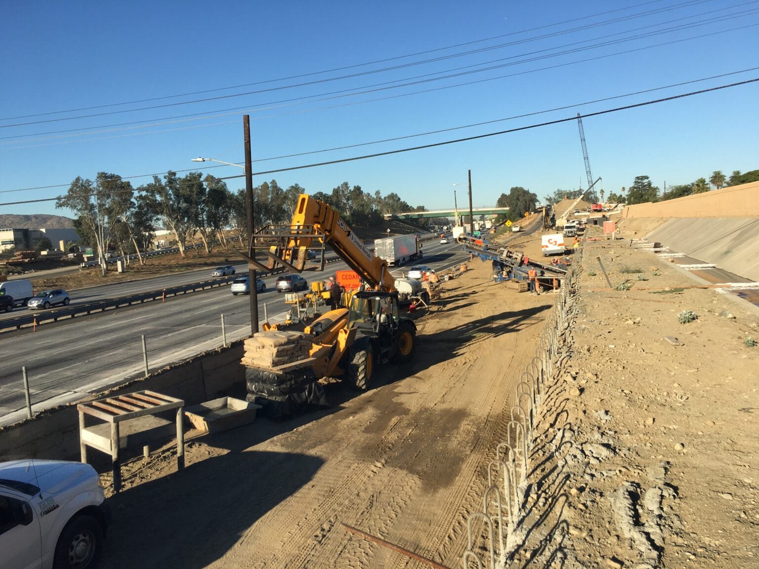 Construction vehicles and workers are present at a highway construction site next to active traffic lanes under a clear sky.