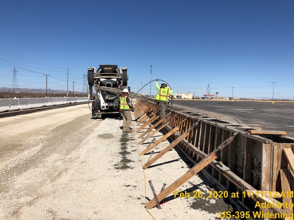 Construction workers pour concrete into a wooden form along a road under clear sky; timestamp and project details are visible in the lower right corner.