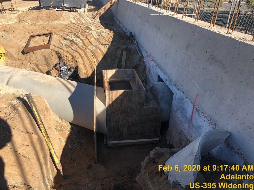 Construction site with exposed drainage pipes, wooden forms, and dirt excavation beside a concrete barrier. Text overlay indicates location and date: Adelanto, US-395 Widening, Feb 6, 2020.