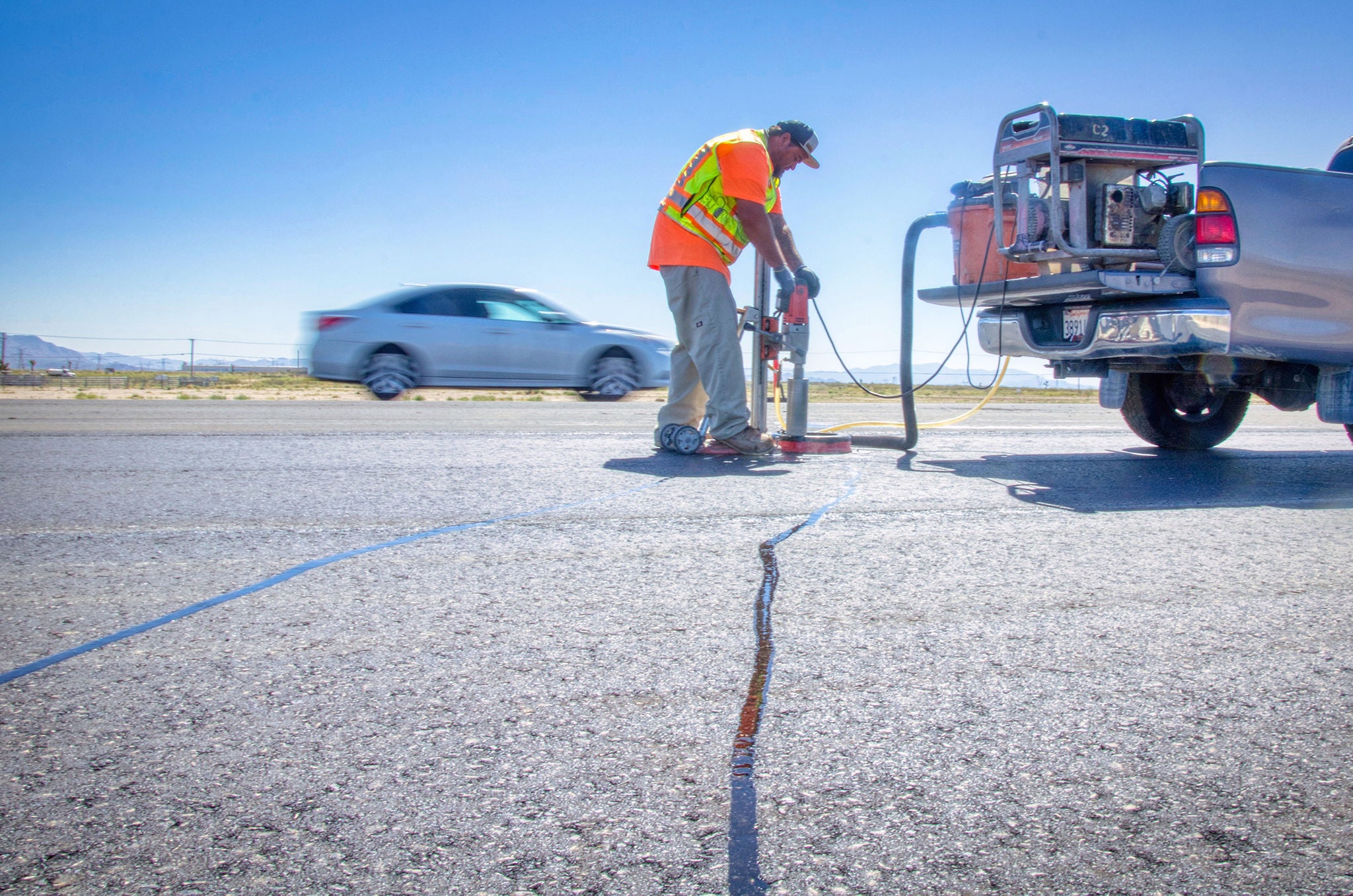 A worker in a safety vest repairs a long crack in a road using equipment attached to a truck, while cars drive by in the background.
