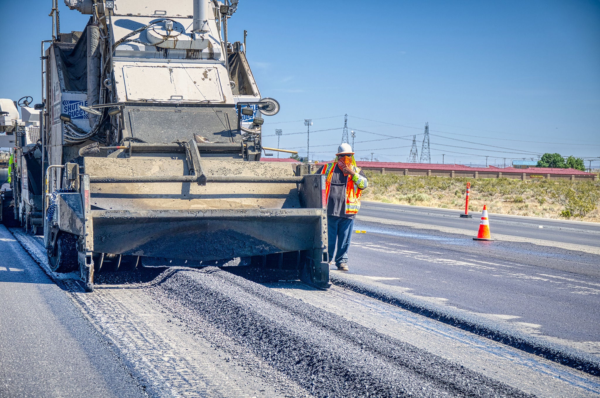 A worker in a safety vest and helmet stands next to a large road paving machine laying asphalt on a multi-lane highway. Traffic cones and power lines are visible in the background.