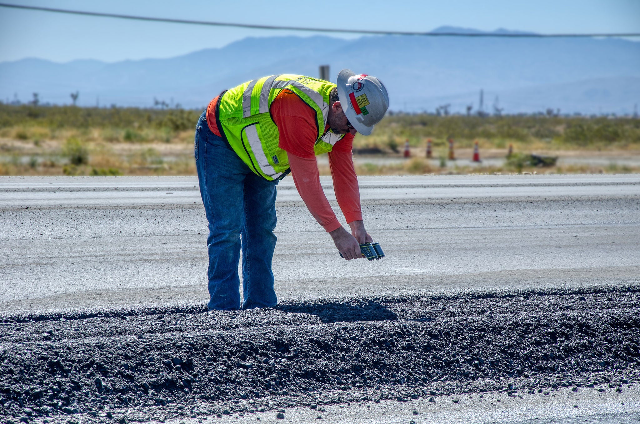 A construction worker in a safety vest and helmet takes a photo of freshly laid asphalt on a road under clear skies.