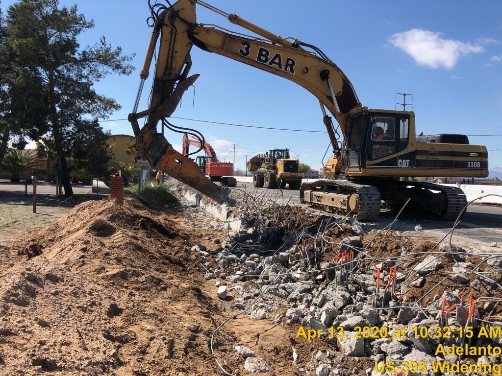 An excavator breaks up concrete and rebar at a construction site next to a dirt road, with construction vehicles and equipment in the background.