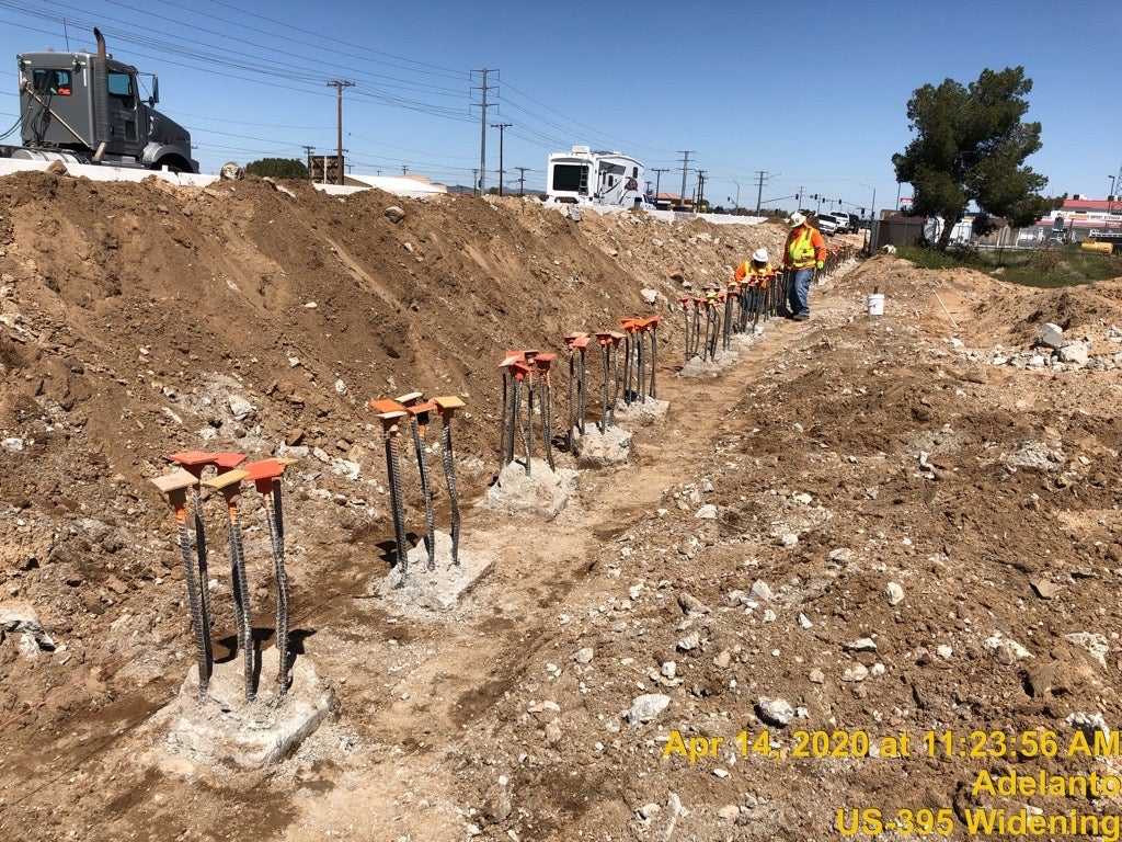 Several construction workers stand in a dirt trench with rows of rebar posts topped with orange safety caps; machinery and vehicles are visible in the background.