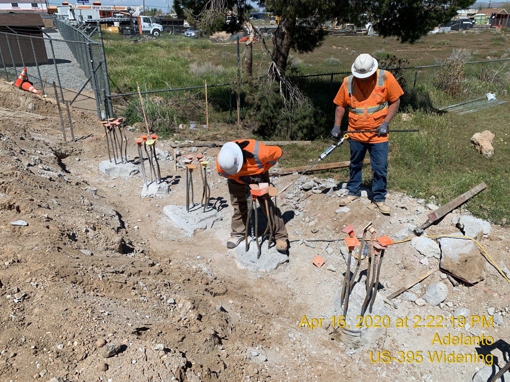 Two construction workers in safety gear inspect and work on rebar rods at a roadside construction site. Date and project details are displayed on the image.