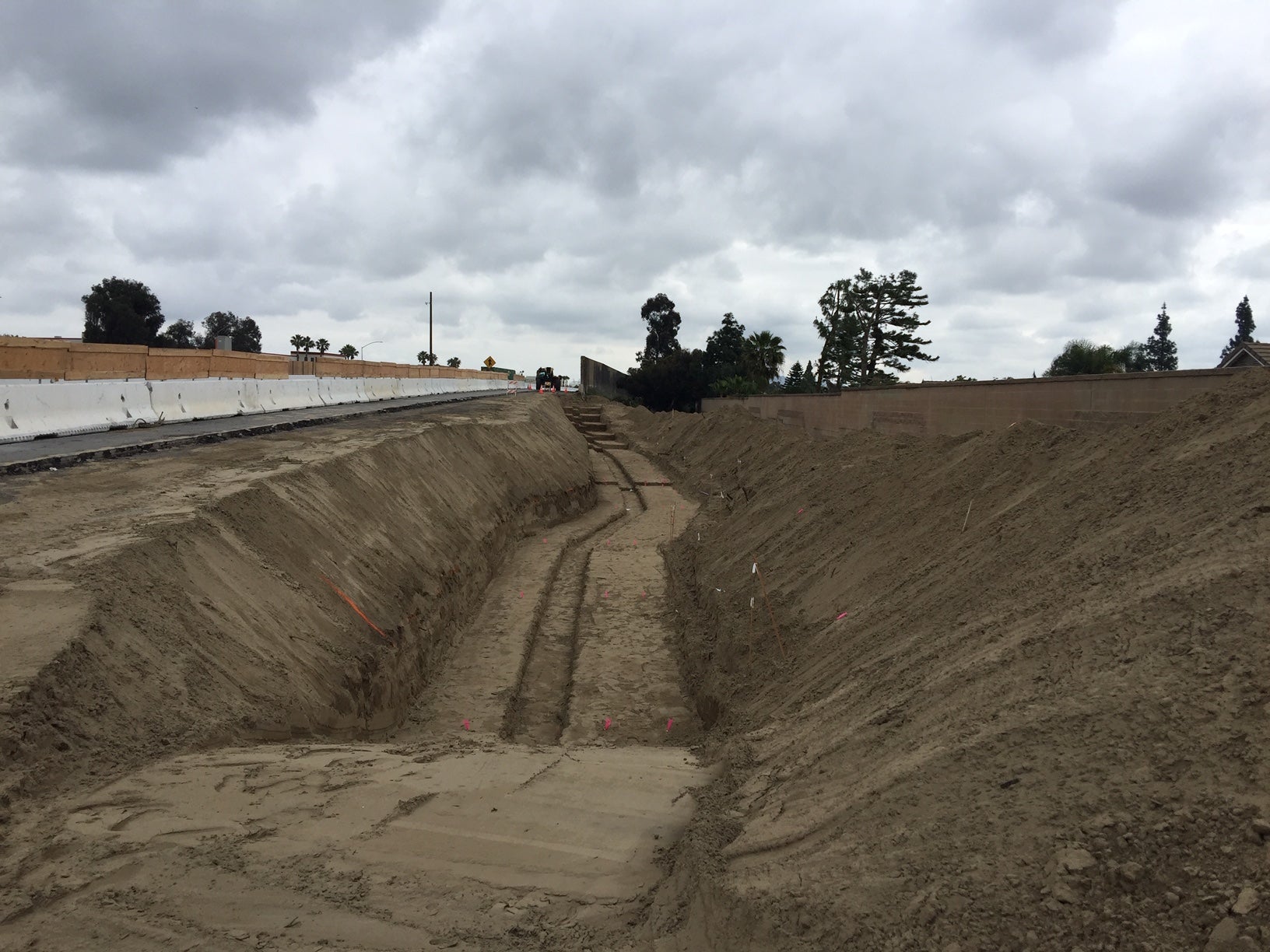 A wide, deep trench runs parallel to a road under cloudy skies, with construction barriers and machinery visible in the background.