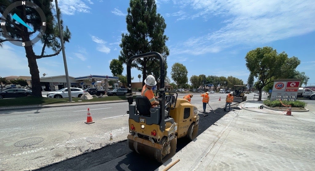 Construction workers pave a section of road with asphalt next to a busy street, using construction equipment and wearing safety gear.