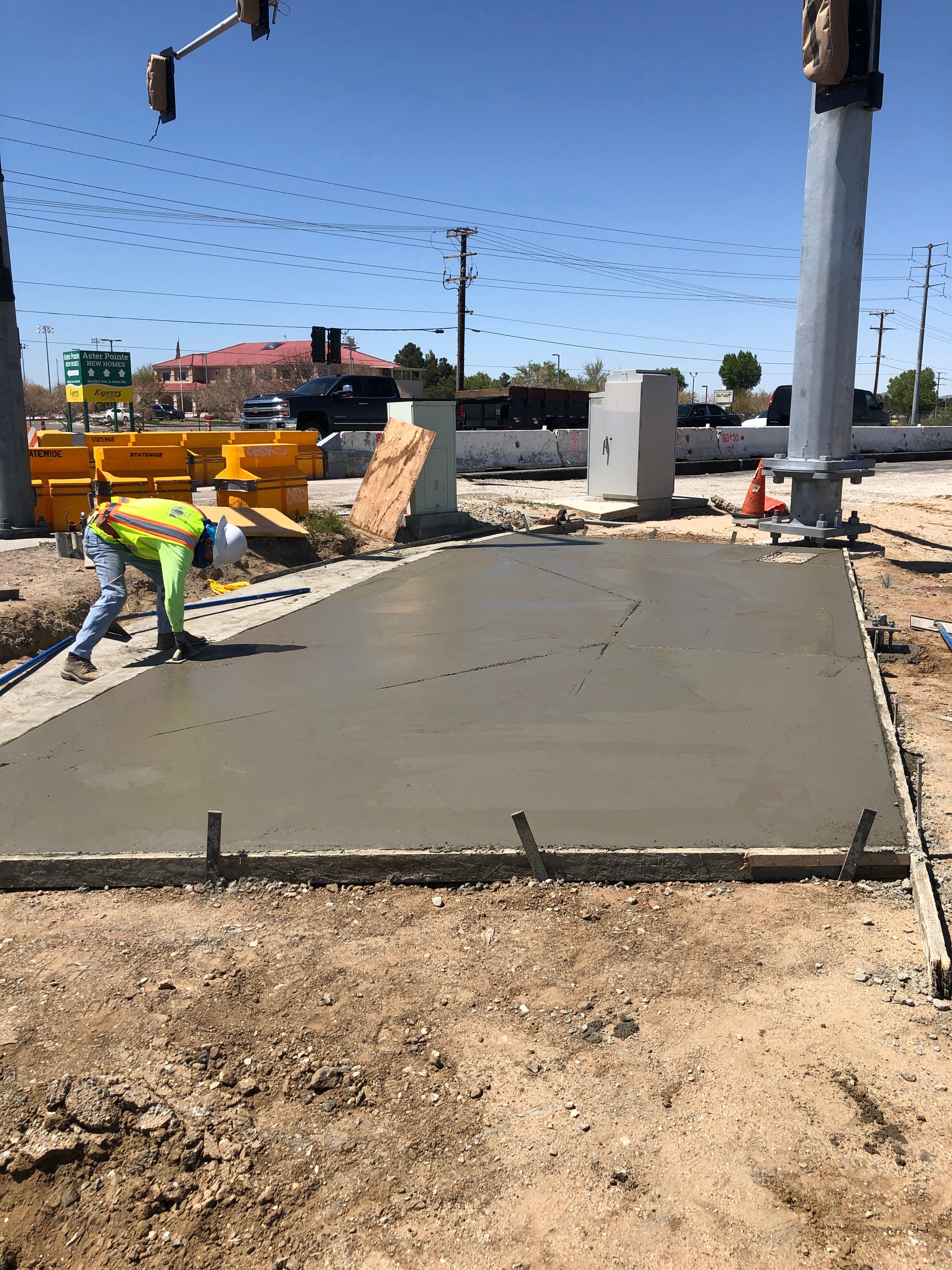 A construction worker smooths wet concrete on a newly poured sidewalk at a road intersection under clear skies.