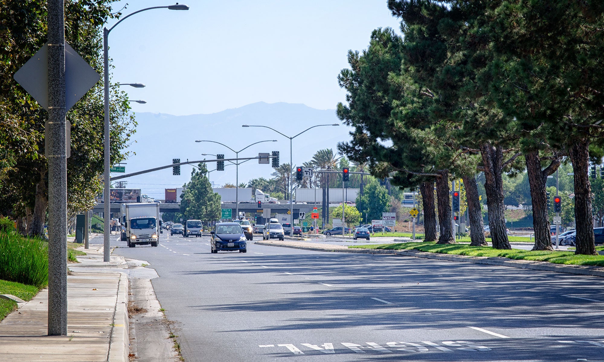 A city street with cars driving in both directions, traffic lights ahead, trees lining the roadway, and mountains visible in the background.