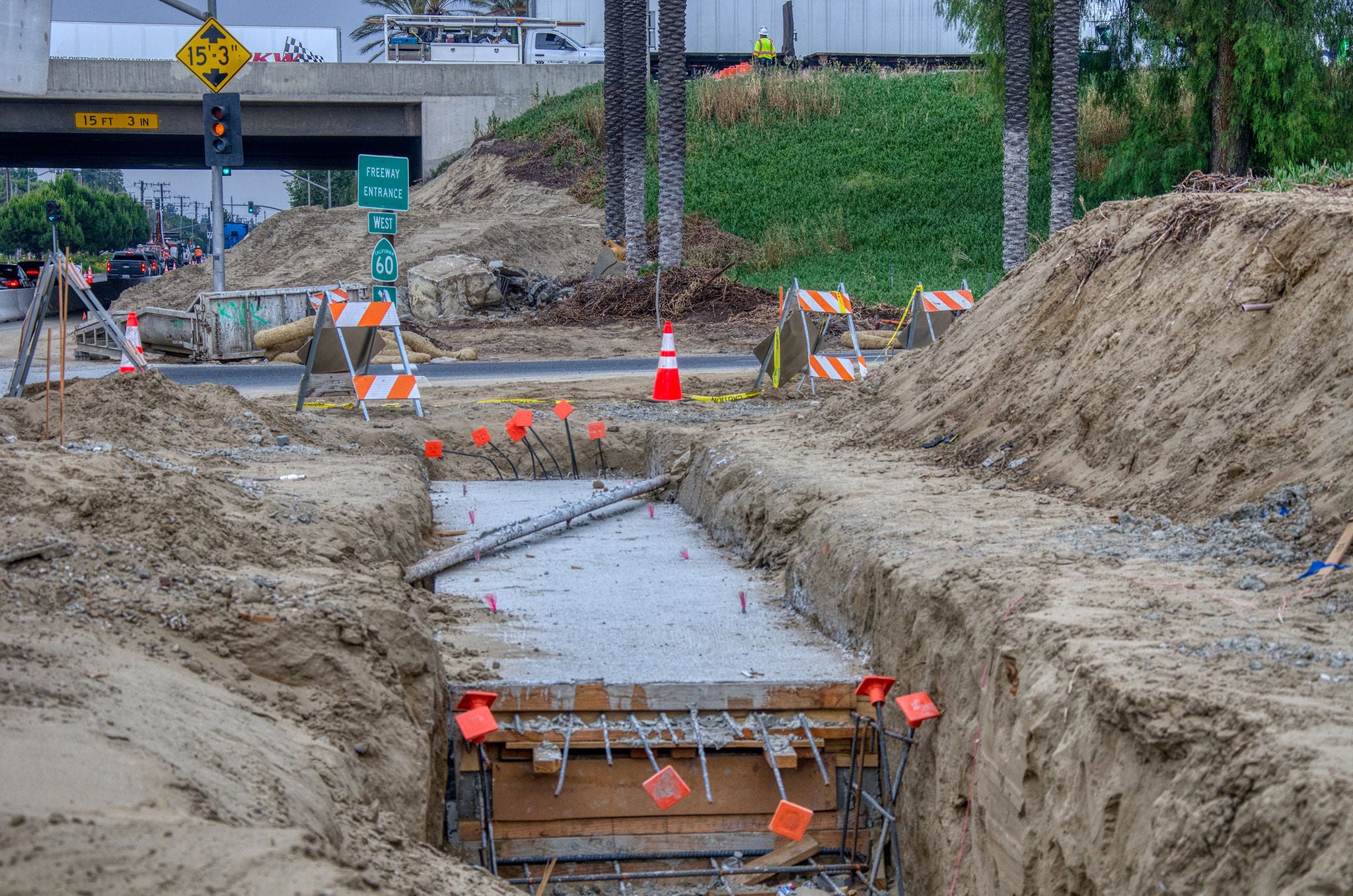 A construction site with a dug trench, traffic barricades, and equipment near a road and overpass. Orange flags and cones mark the area for safety.