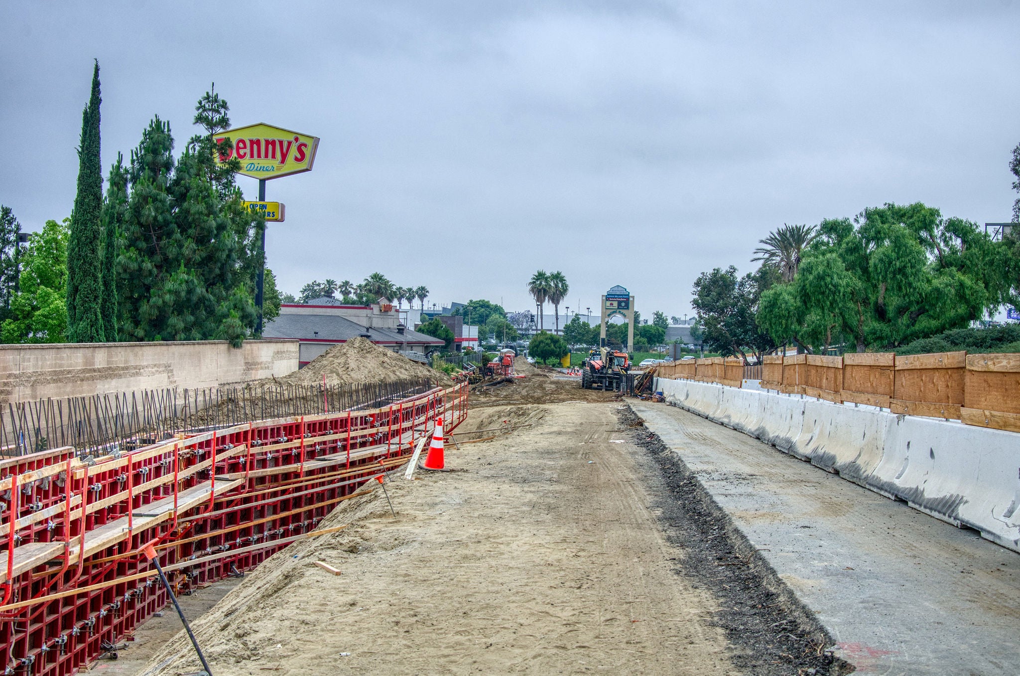A partially constructed road with construction barriers, equipment, and dirt piles; a Denny’s restaurant sign is visible in the background on a cloudy day.