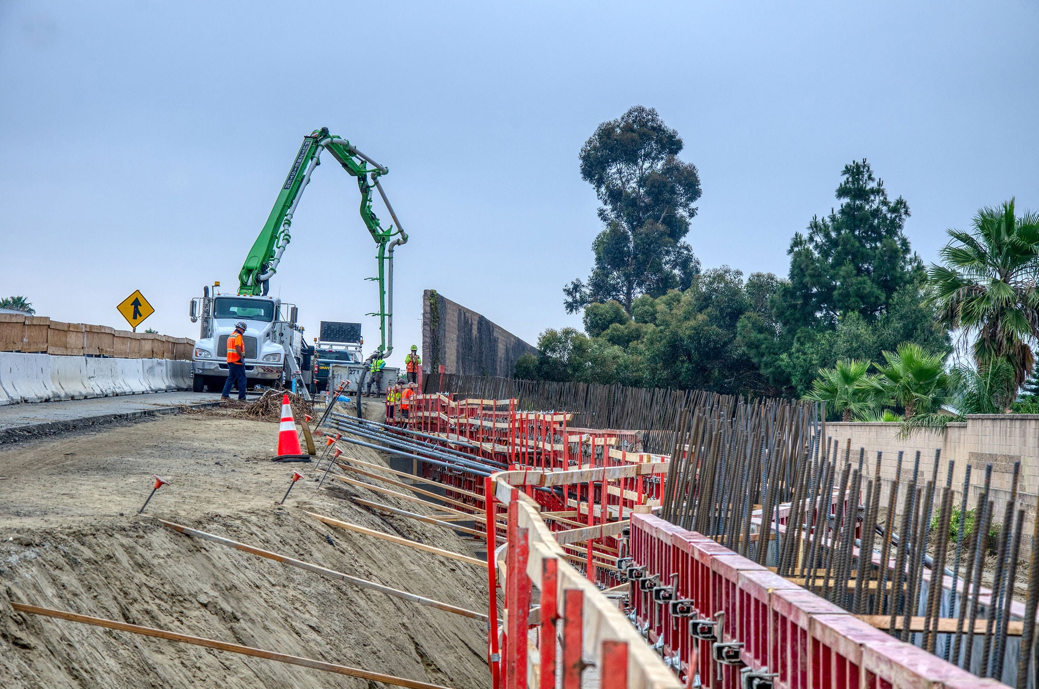 Construction workers and machinery are building a concrete structure alongside a road, with barriers, rebar, and trees in the background.