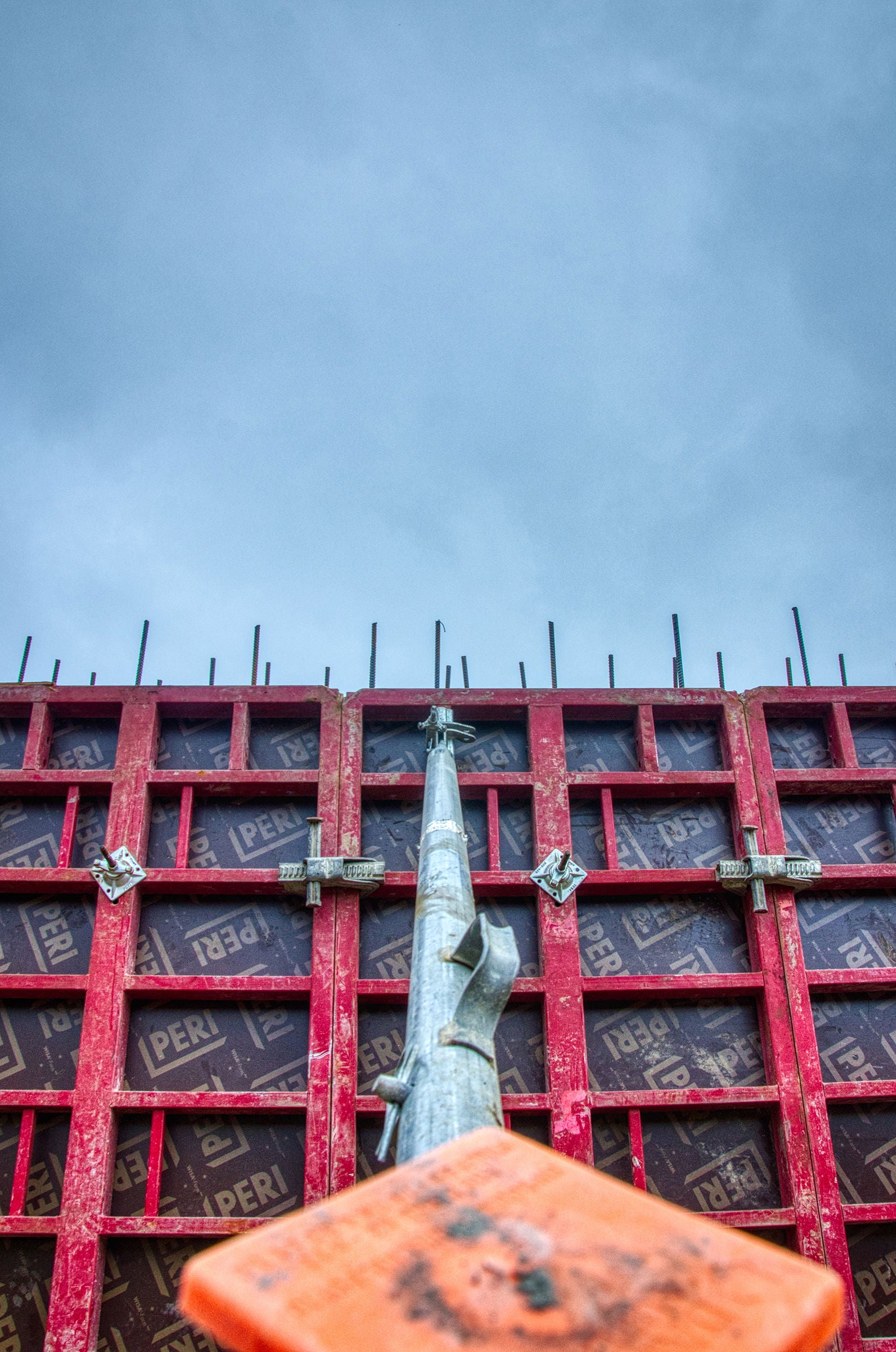 A red construction formwork structure with exposed rebar is viewed from below, with a metal support pole extending upward toward a cloudy sky.