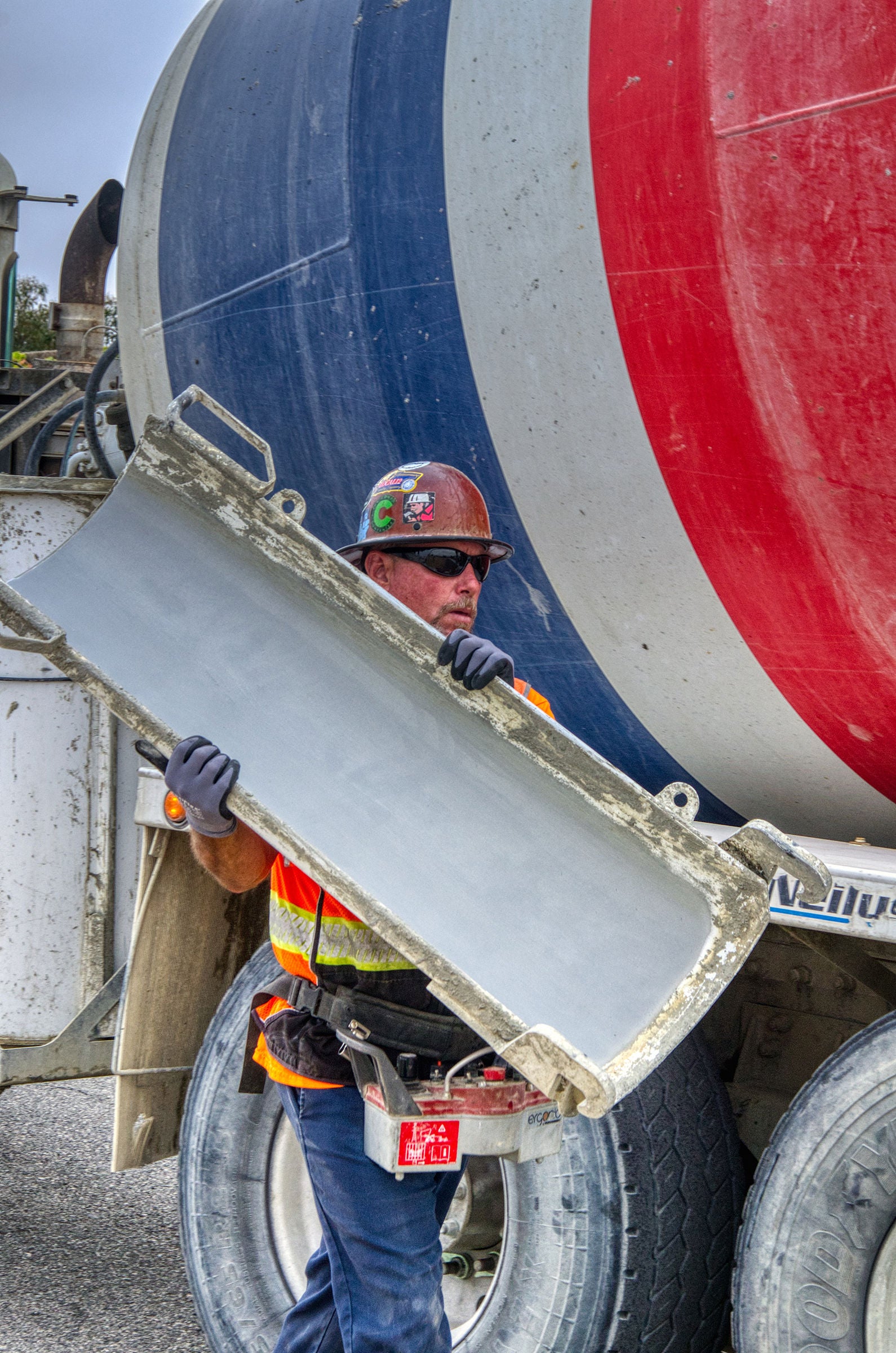 A construction worker wearing safety gear holds a concrete chute in front of a cement mixer truck with a red, white, and blue drum.