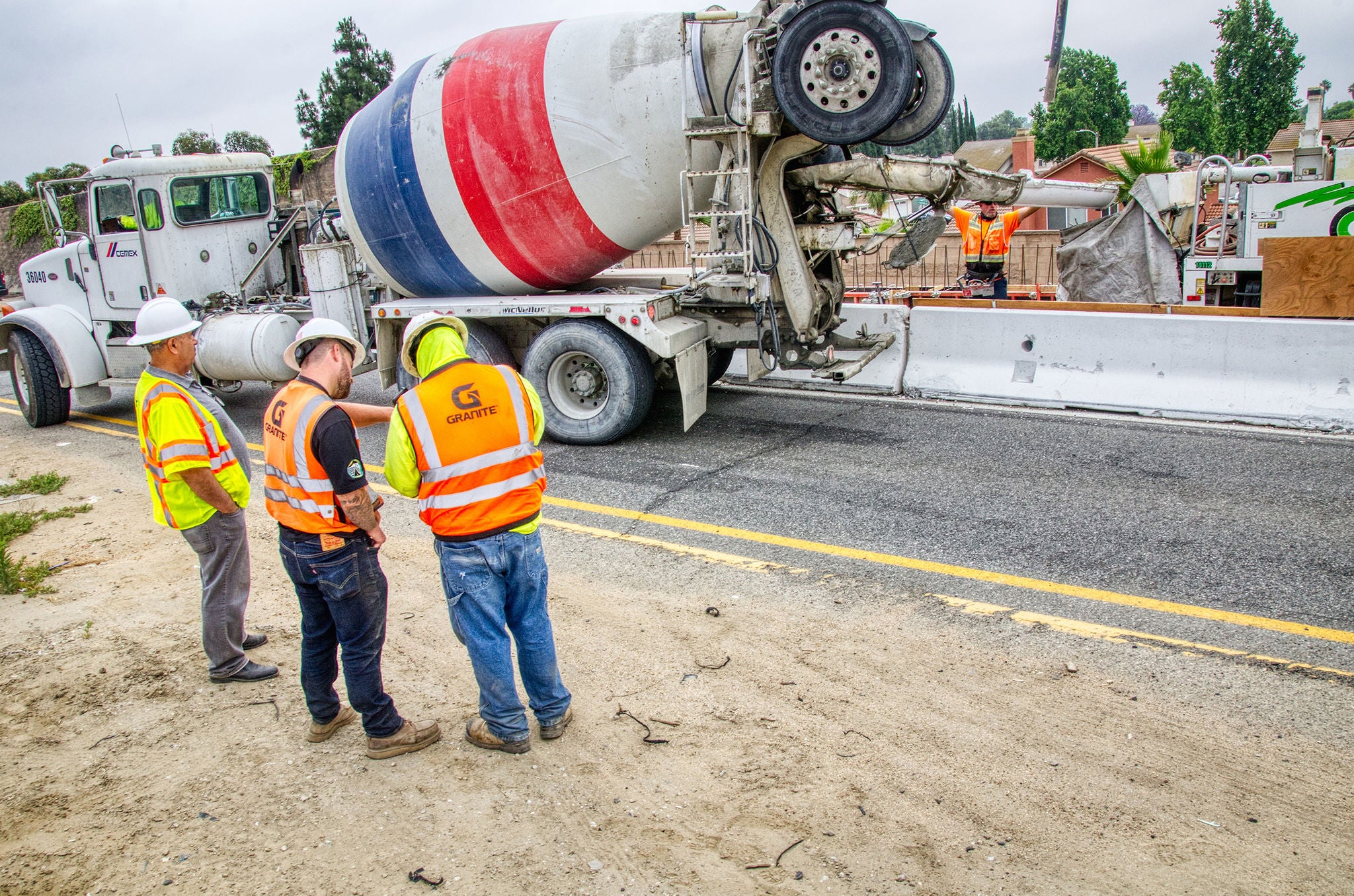 Three construction workers in safety gear stand near a cement mixer truck at a roadside construction site, while two other workers work behind a barrier.