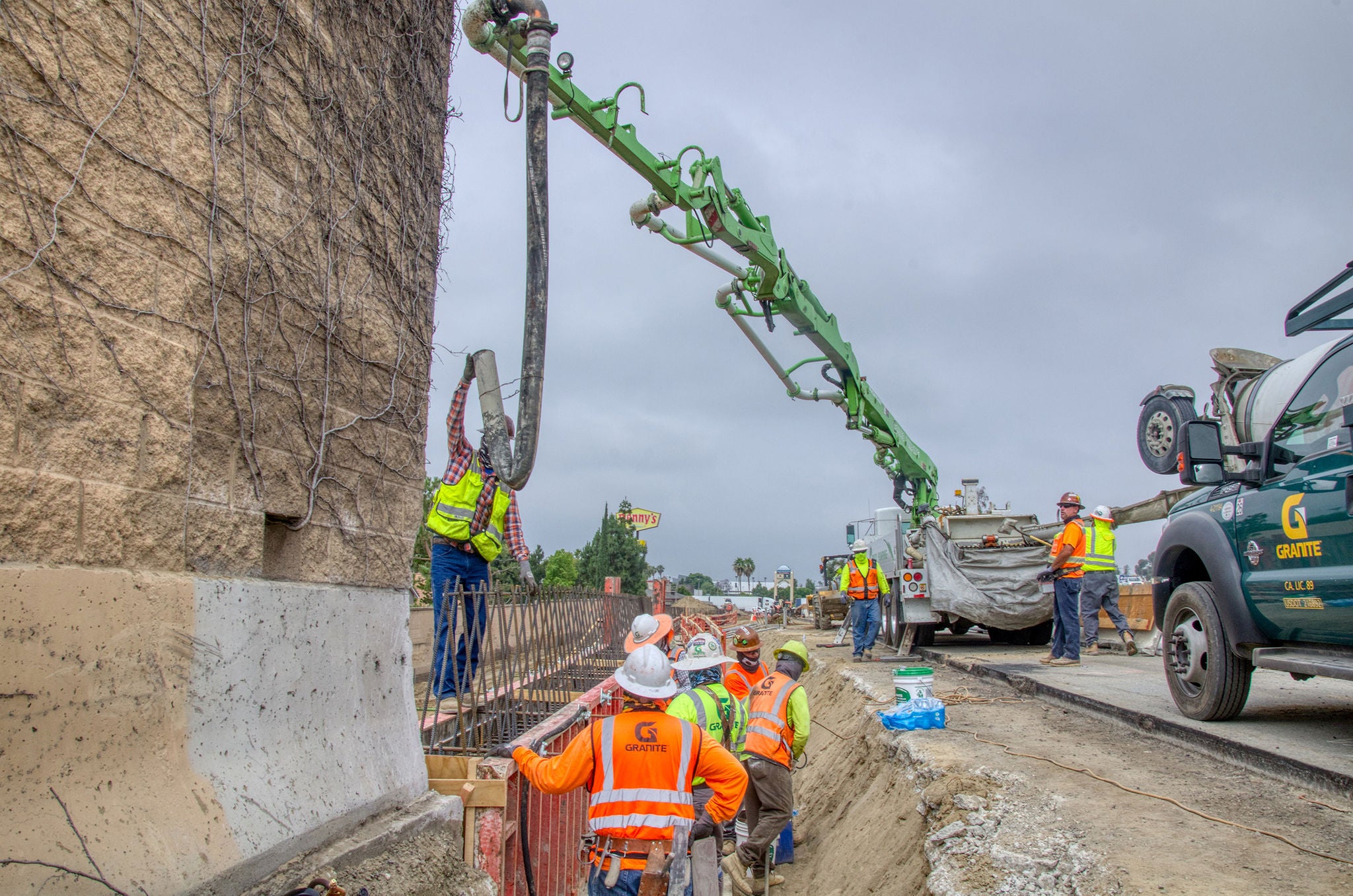 Construction workers oversee and guide a concrete pump hose as it pours concrete into a form next to a busy roadway.
