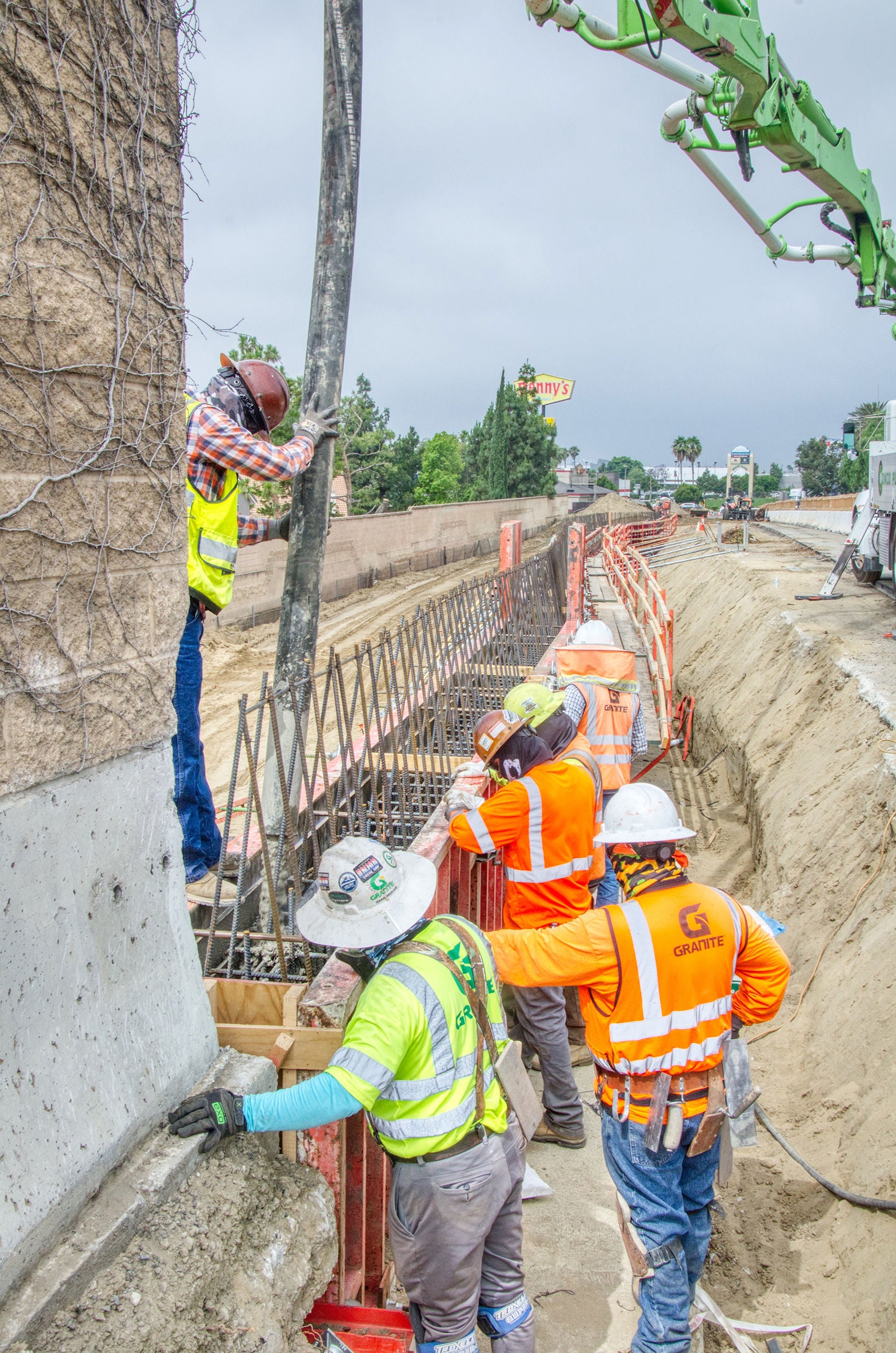 Construction workers in safety gear pour concrete into a foundation near a busy road, using a large hose and reinforced steel rebar for support.