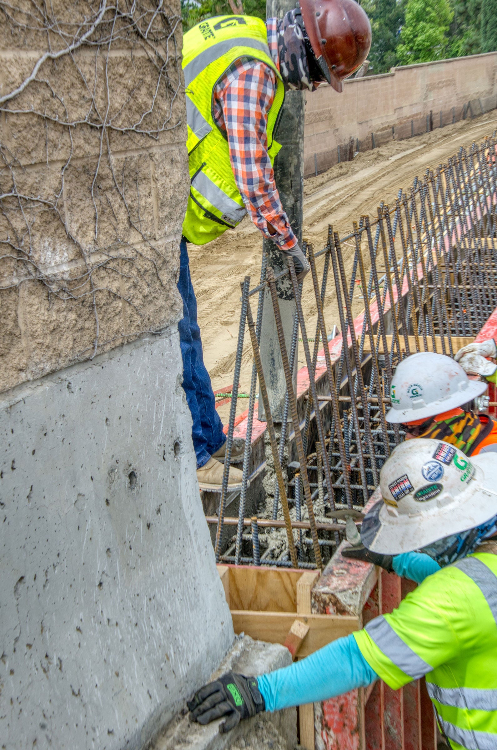 Two construction workers pour concrete into a form reinforced with rebar at a construction site; both are wearing safety gear including helmets and vests.