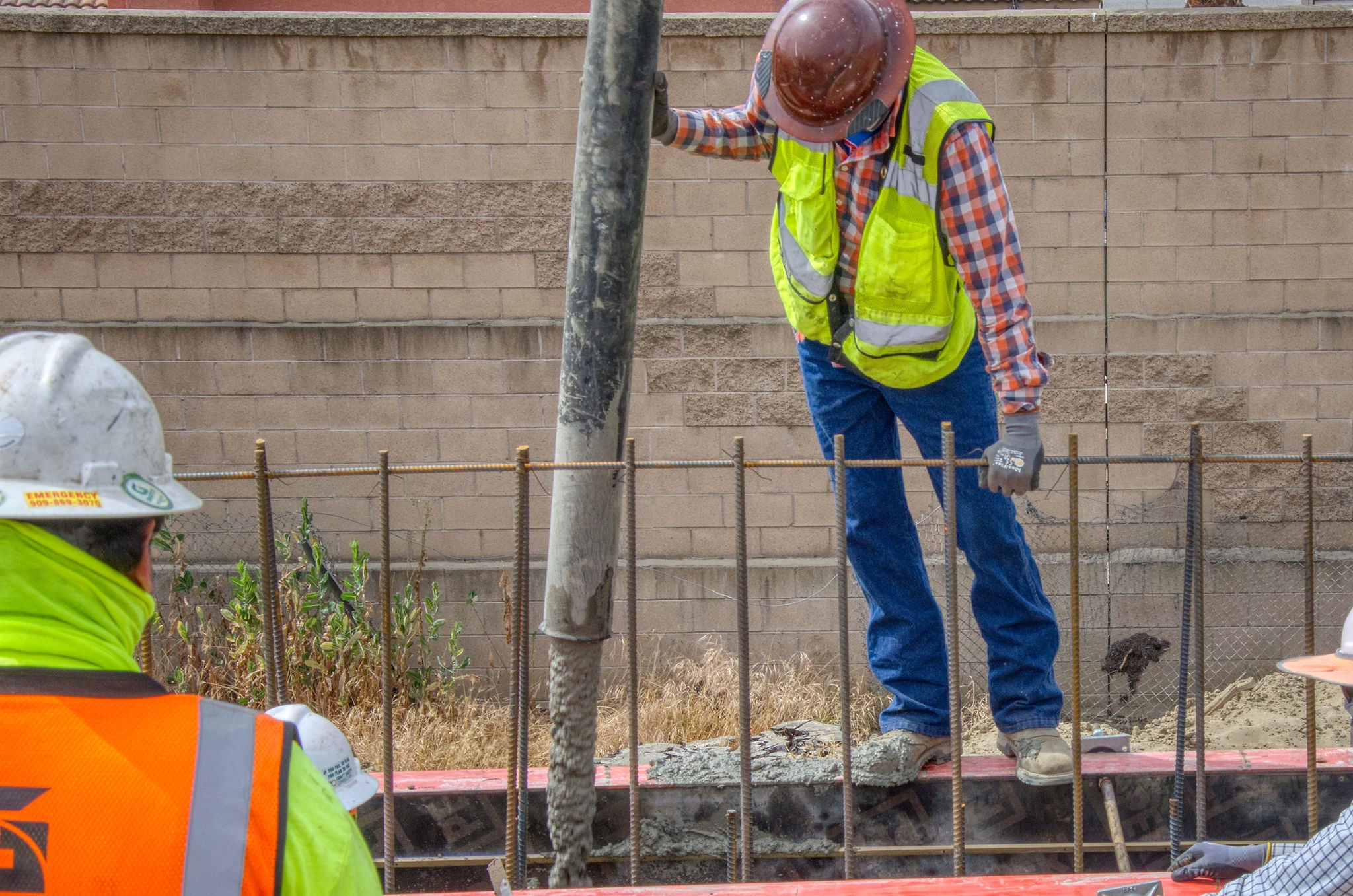 Construction worker in a safety vest and helmet guides a concrete hose while standing on a rebar-framed section of a building site.