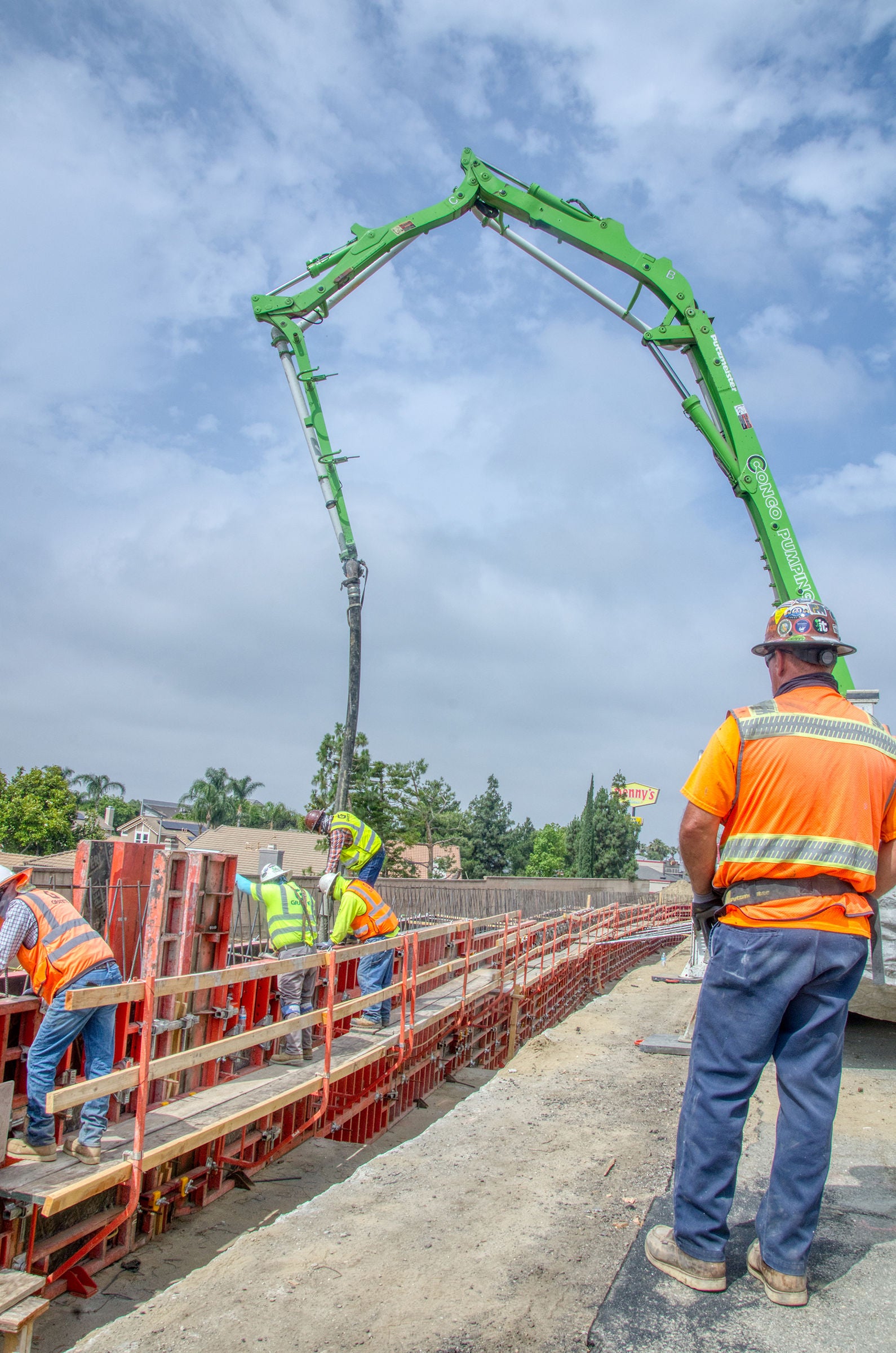 Construction workers operate a large green concrete pump, pouring concrete into a formwork structure at an outdoor construction site.