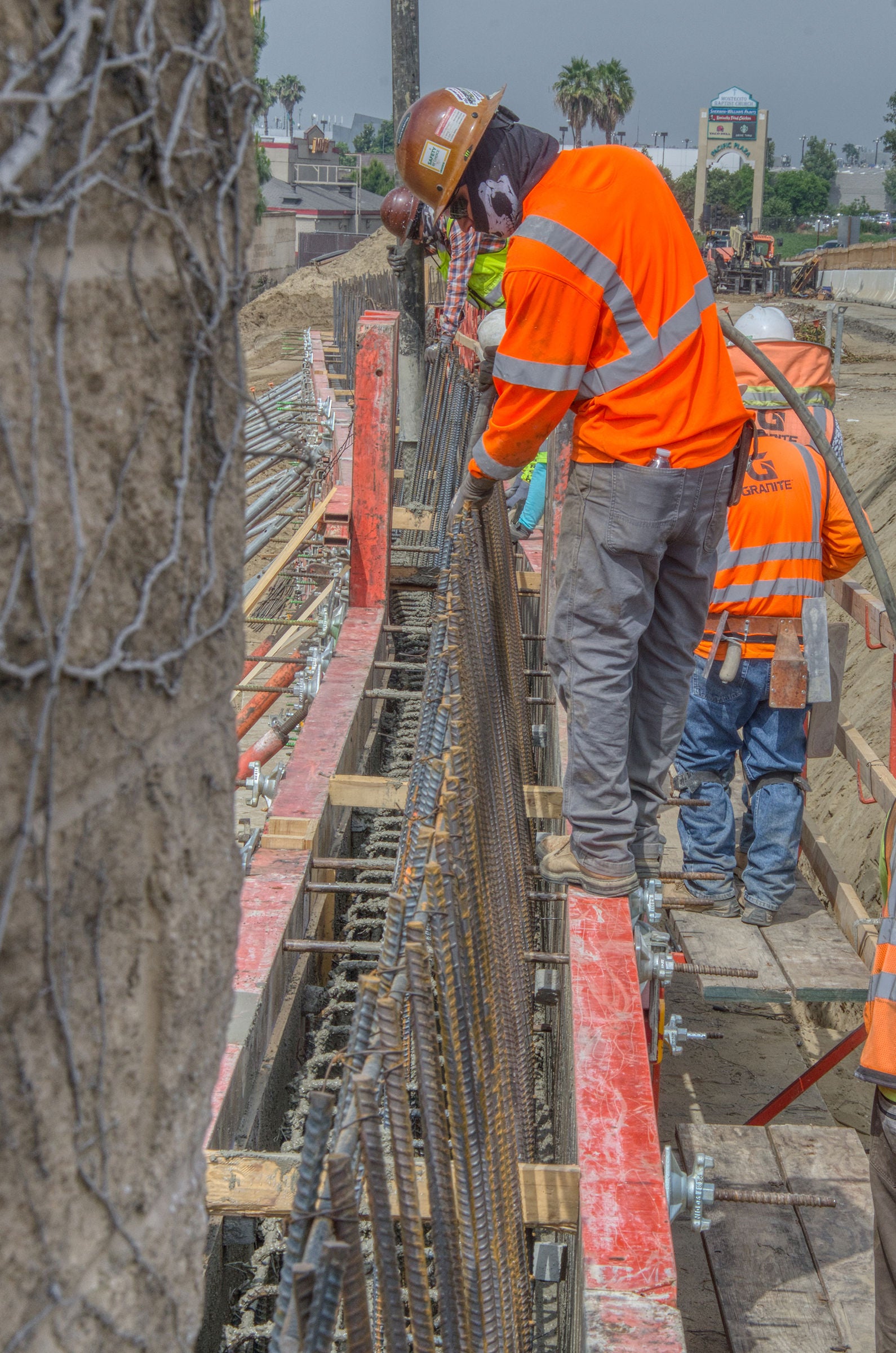 Construction workers in orange safety gear install rebar on a bridge or overpass under construction, using temporary wooden supports on a work platform.