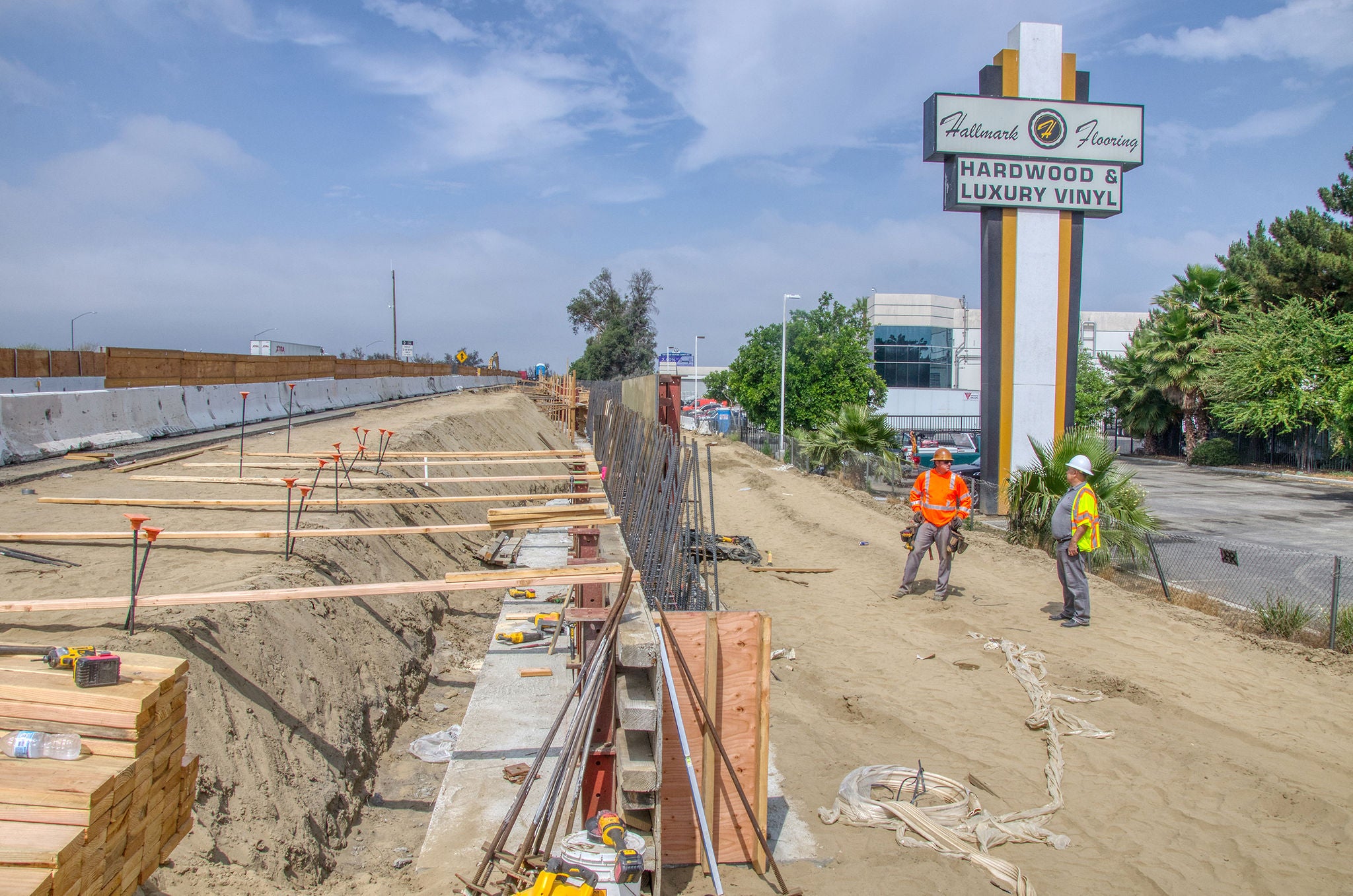 Two construction workers stand near a worksite with wooden beams and tools, adjacent to a "Hardwood & Luxury Vinyl" flooring store sign.