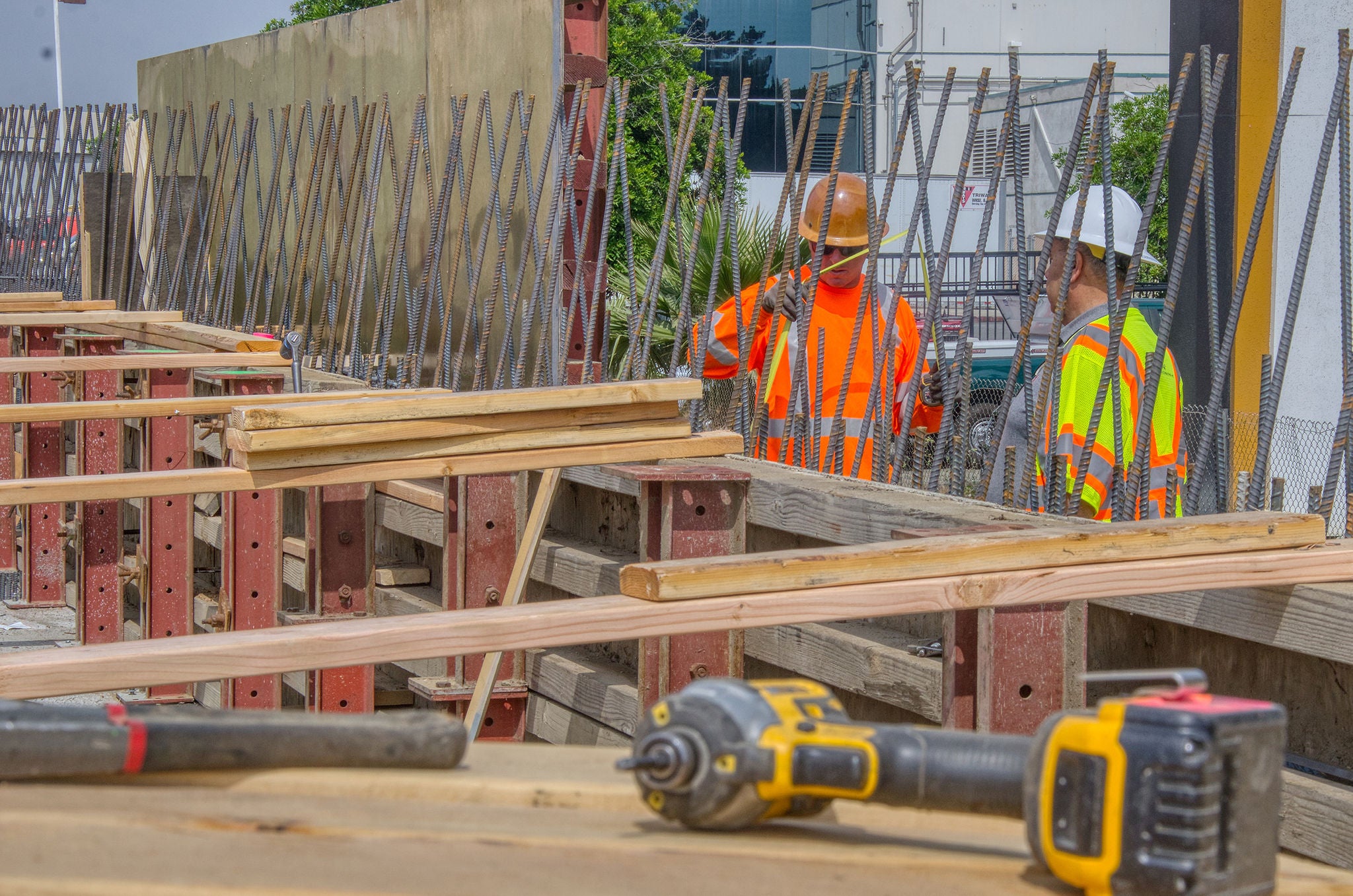 Two construction workers in safety gear stand beside a structure with exposed rebar and wooden planks; various tools are visible in the foreground.