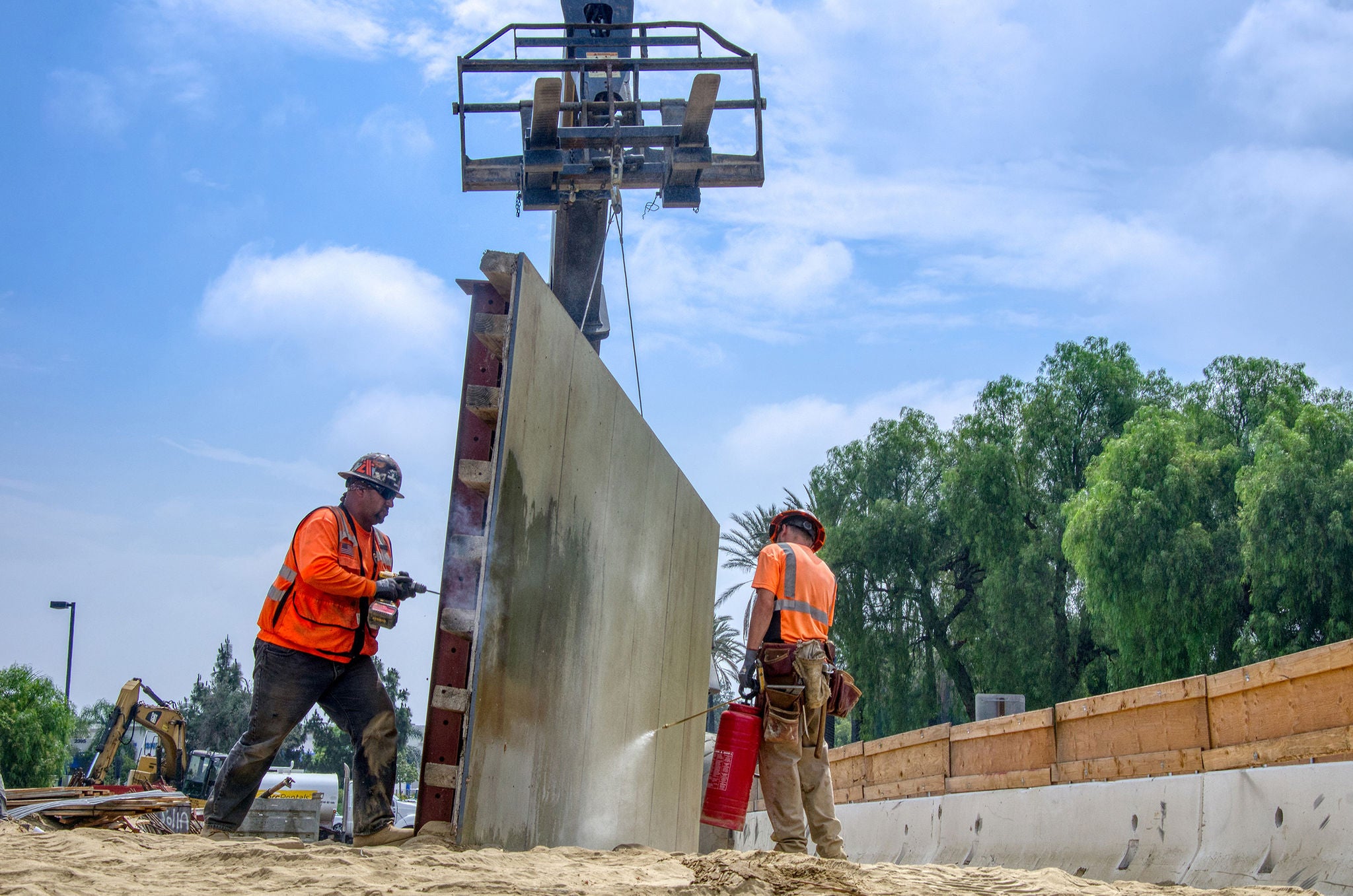 Two construction workers in safety gear guide a large concrete wall panel being positioned by a crane at an outdoor construction site.