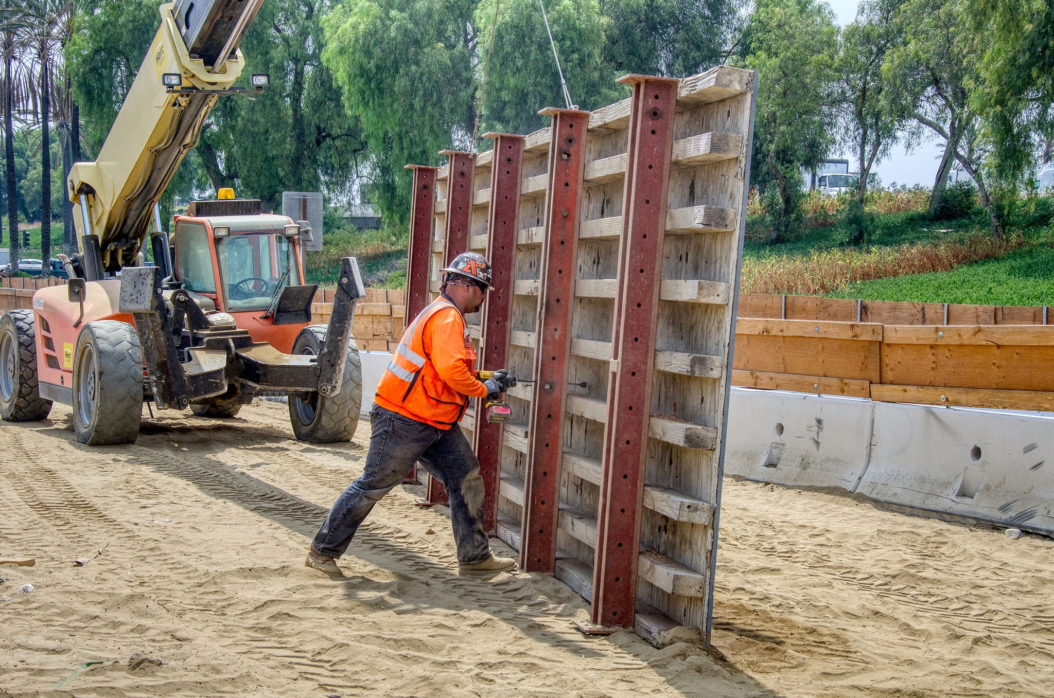 Construction worker in an orange vest and helmet positions a large metal formwork panel at a construction site with a forklift in the background.