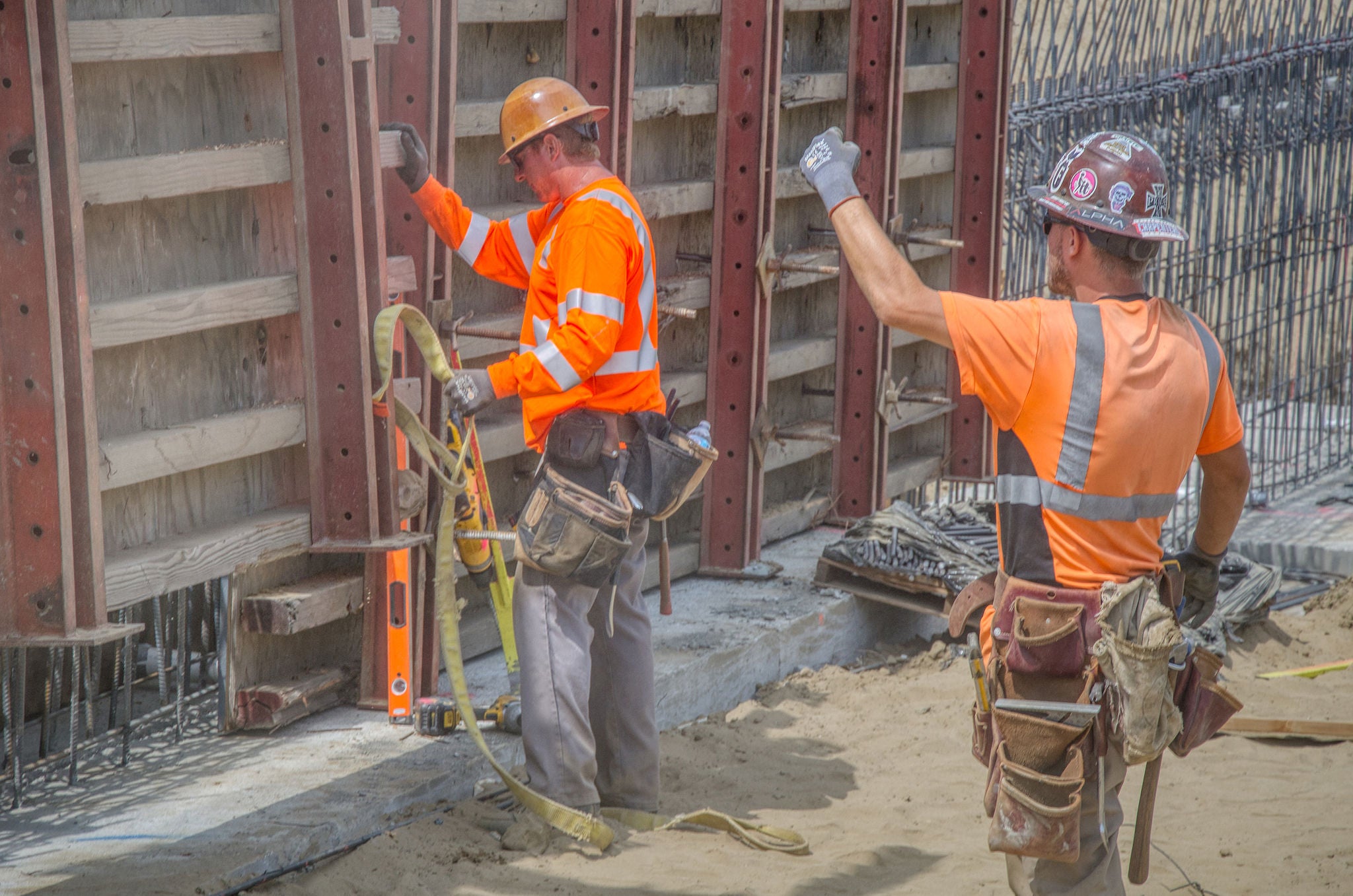 Two construction workers in orange safety gear work with a wall form and leveling tool at a construction site, with one worker raising an arm.