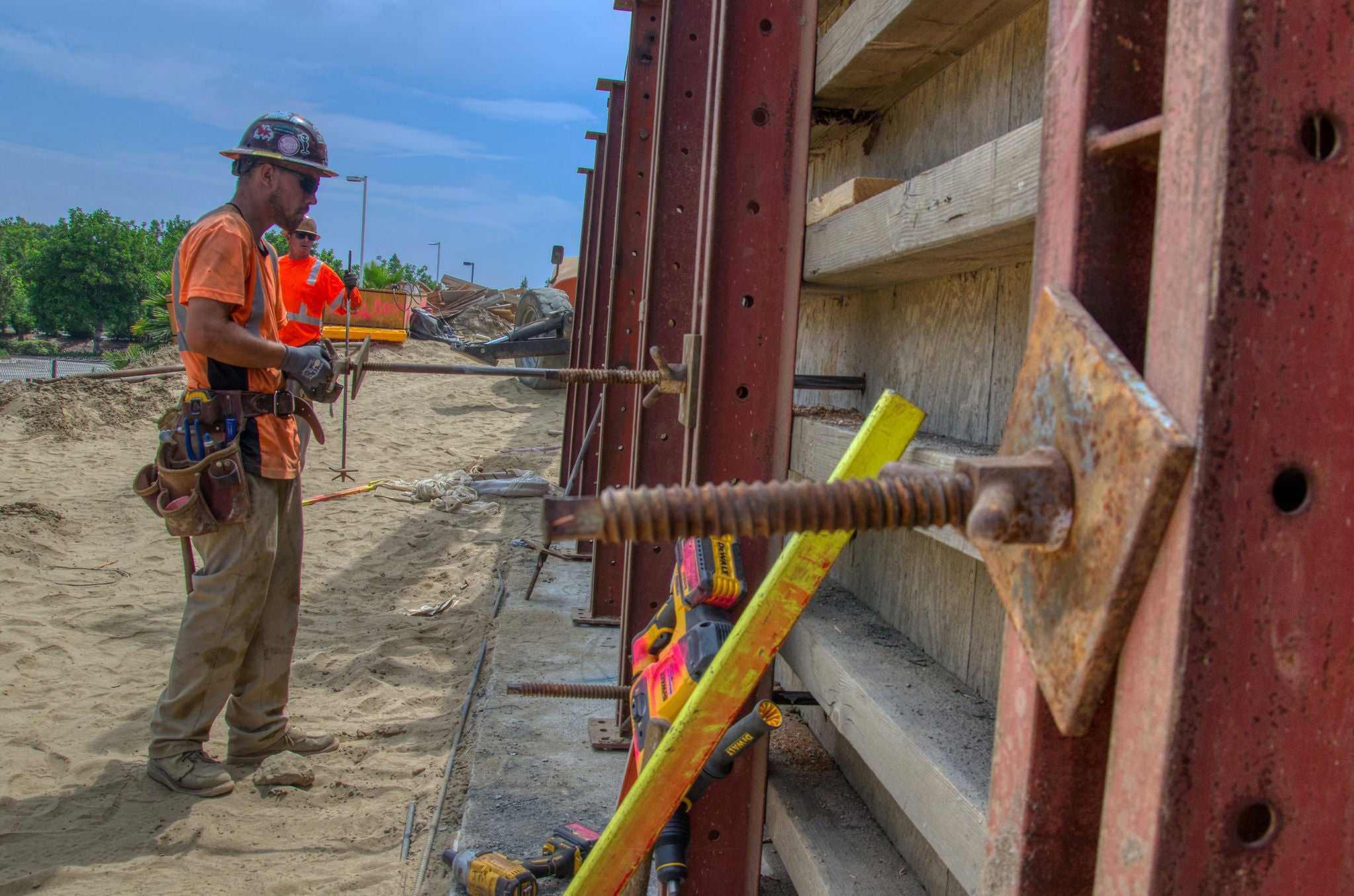 Two construction workers stand by a steel and wood structure at a sandy outdoor work site, with tools and safety gear visible.